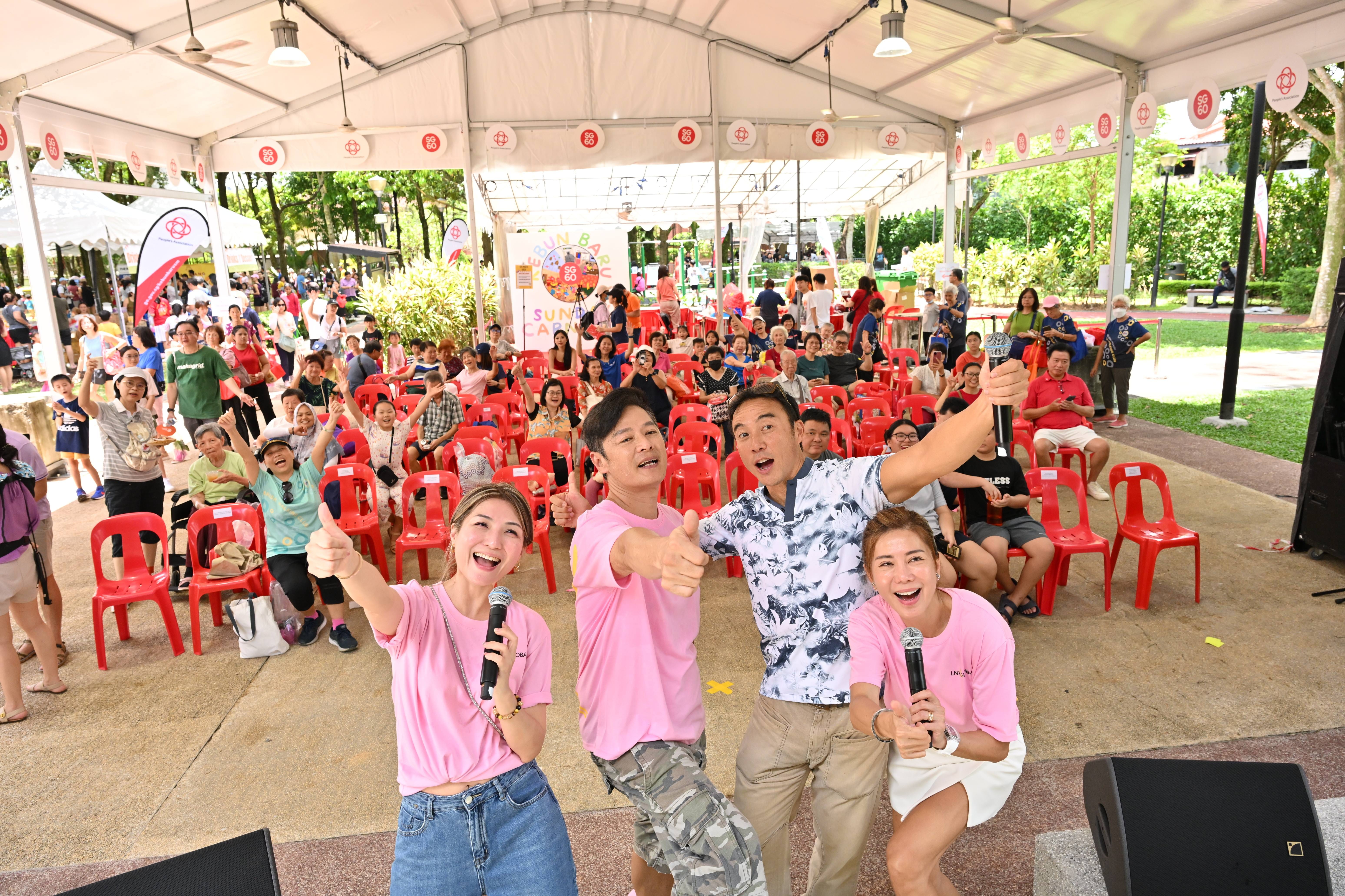 Four smiling emcees in pink shirts hold microphones and give thumbs up before a large seated crowd at an outdoor tent event.