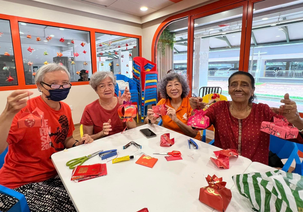 Four elderly women sit at a table displaying handmade red paper crafts, with scissors and materials spread around them.