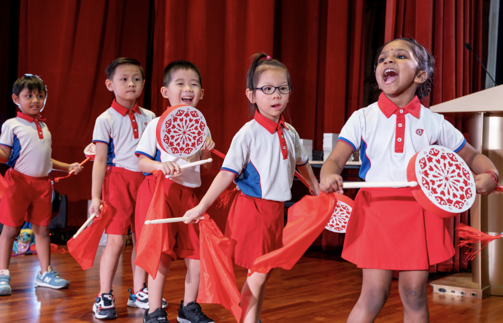 Five young children in red and white uniforms perform a dance holding decorative fans and red scarves on a wooden stage.