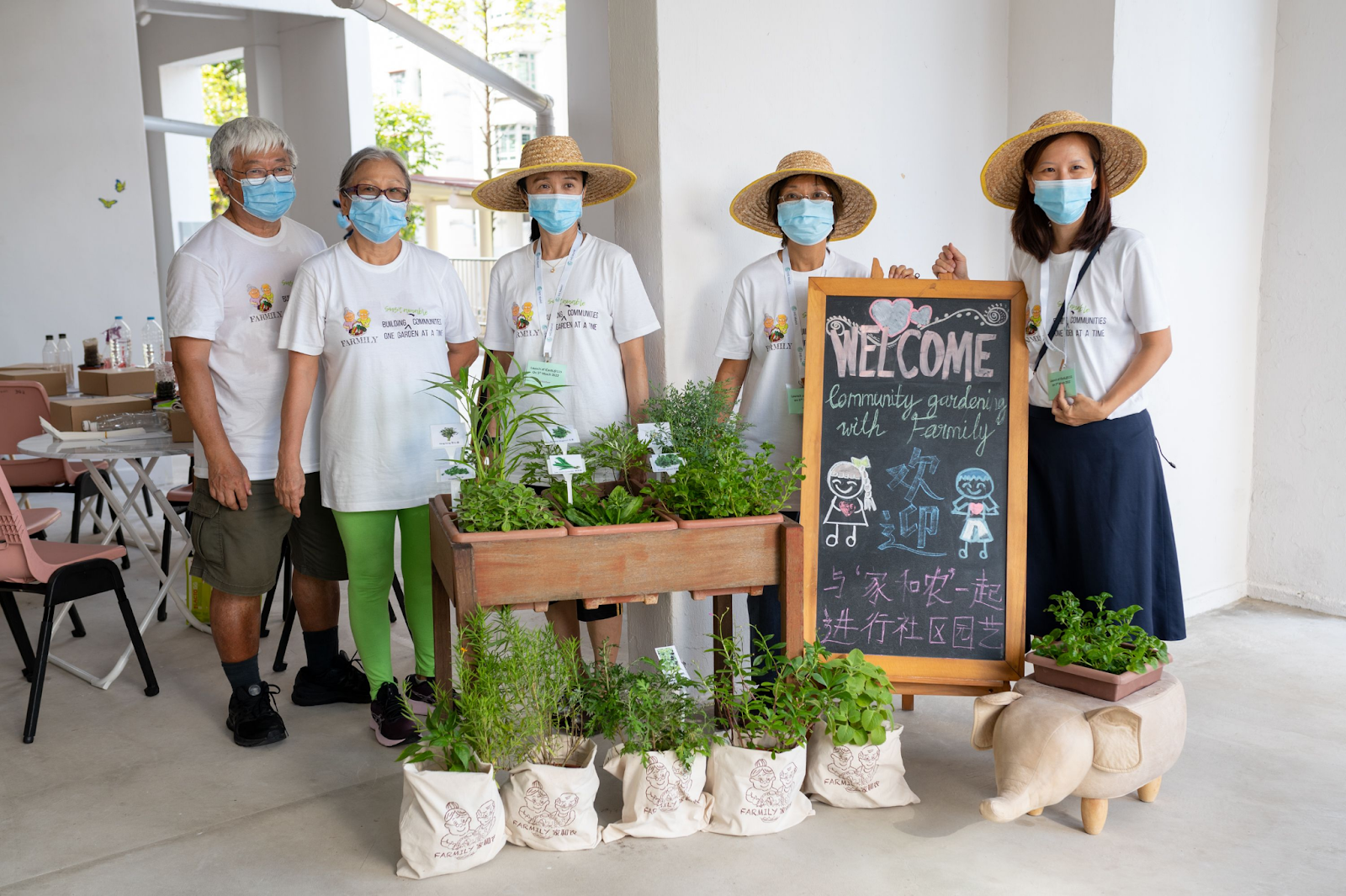 Five masked volunteers in white shirts stand beside potted herbs and a chalkboard reading Welcome Community Gardening with Farmily.