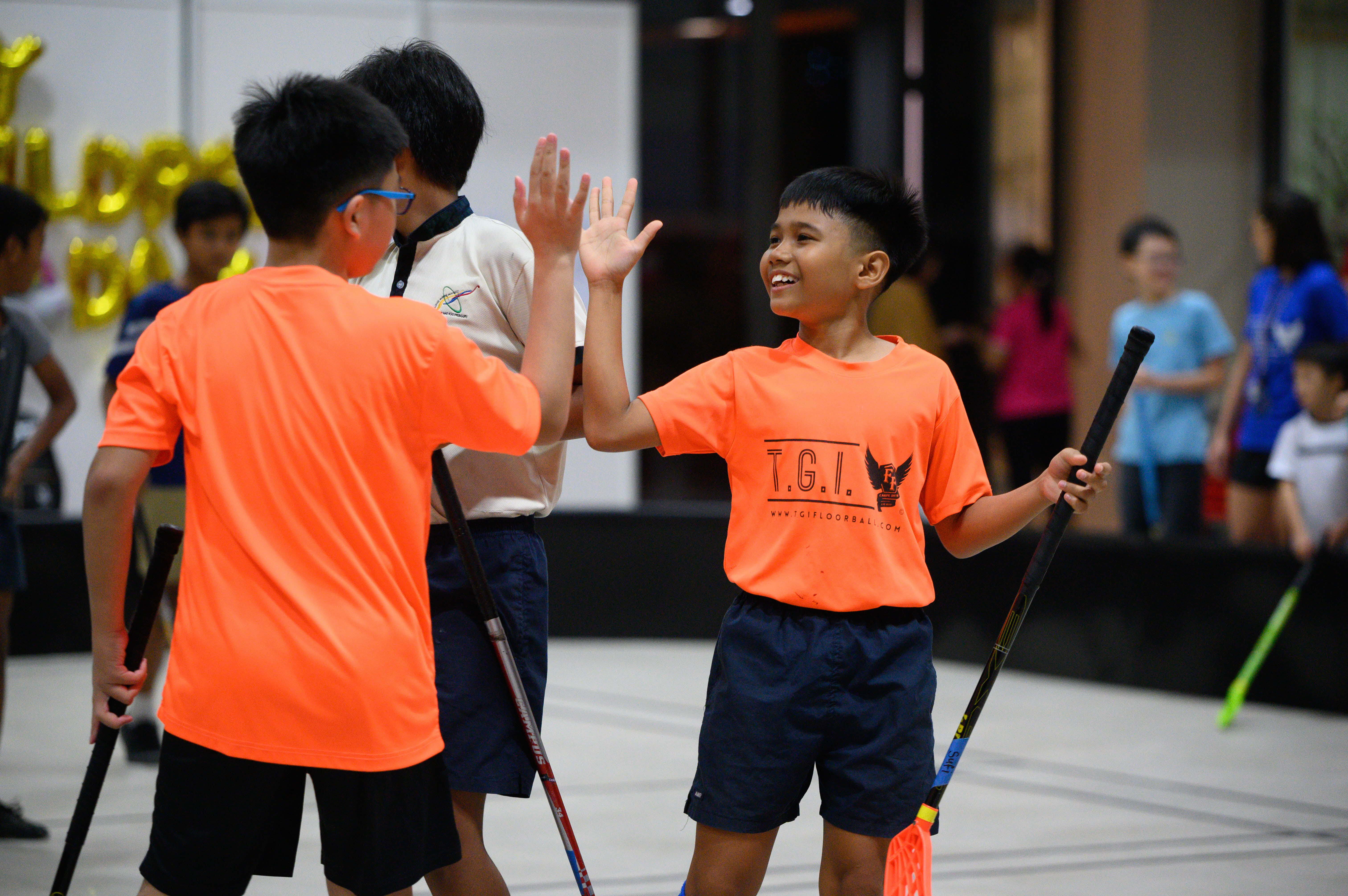 Two boys in orange TGI Floorball shirts high-five while holding sticks during an indoor floorball event.