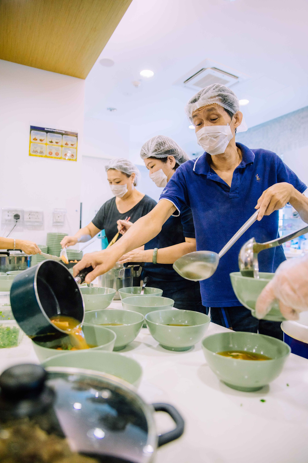 Three masked workers in hairnets ladle soup broth into rows of green bowls in a commercial kitchen.
