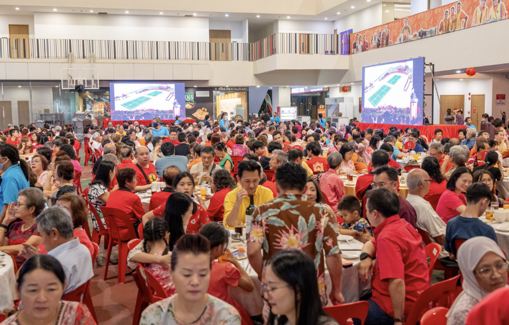Large indoor banquet hall packed with people seated at round tables, many wearing red, with two large screens at the front.