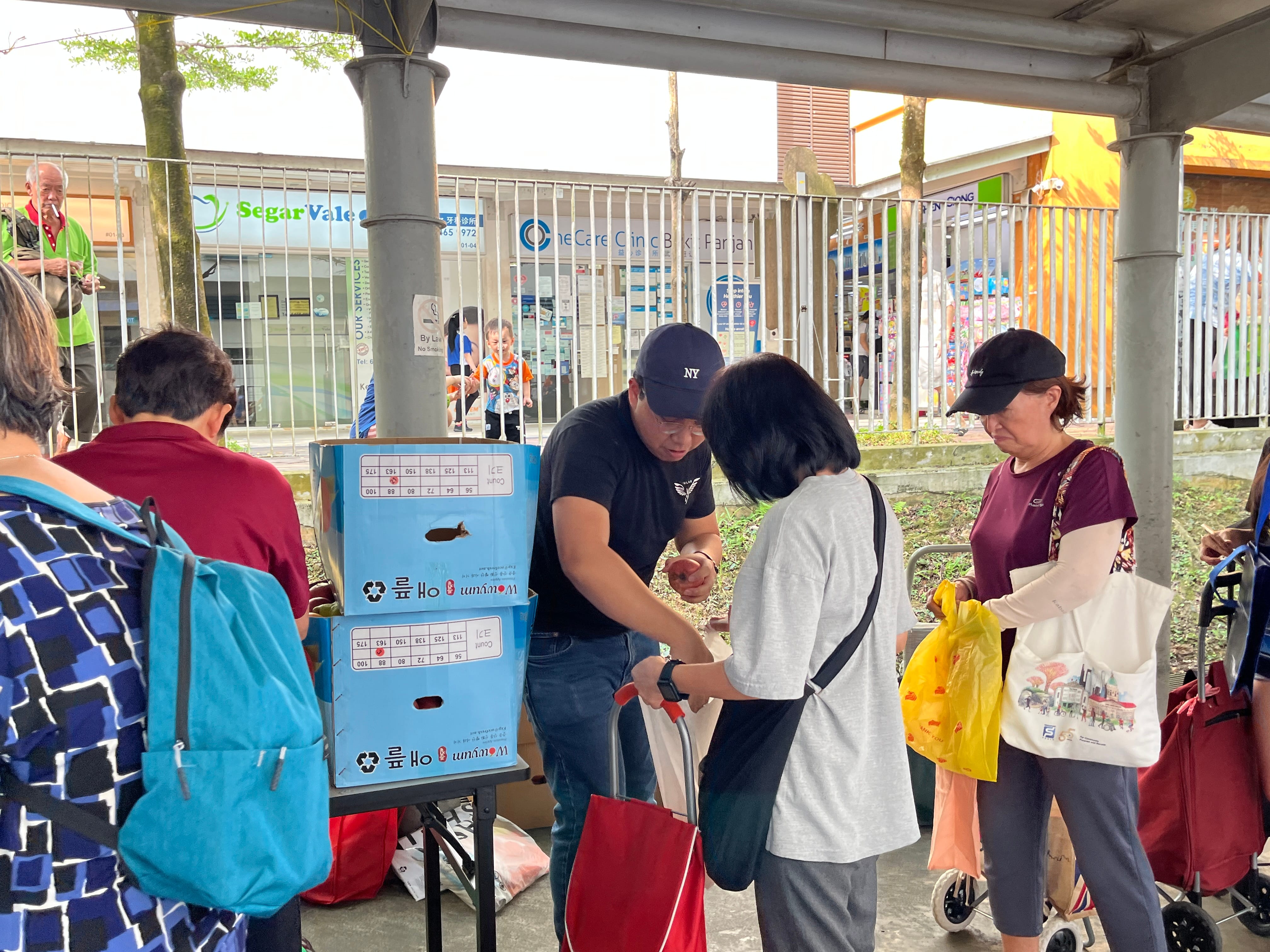 Several people gather under a covered walkway near blue cardboard boxes. A volunteer male wearing a black tshirt, jeans and a dark blue cap is distributing fruits.