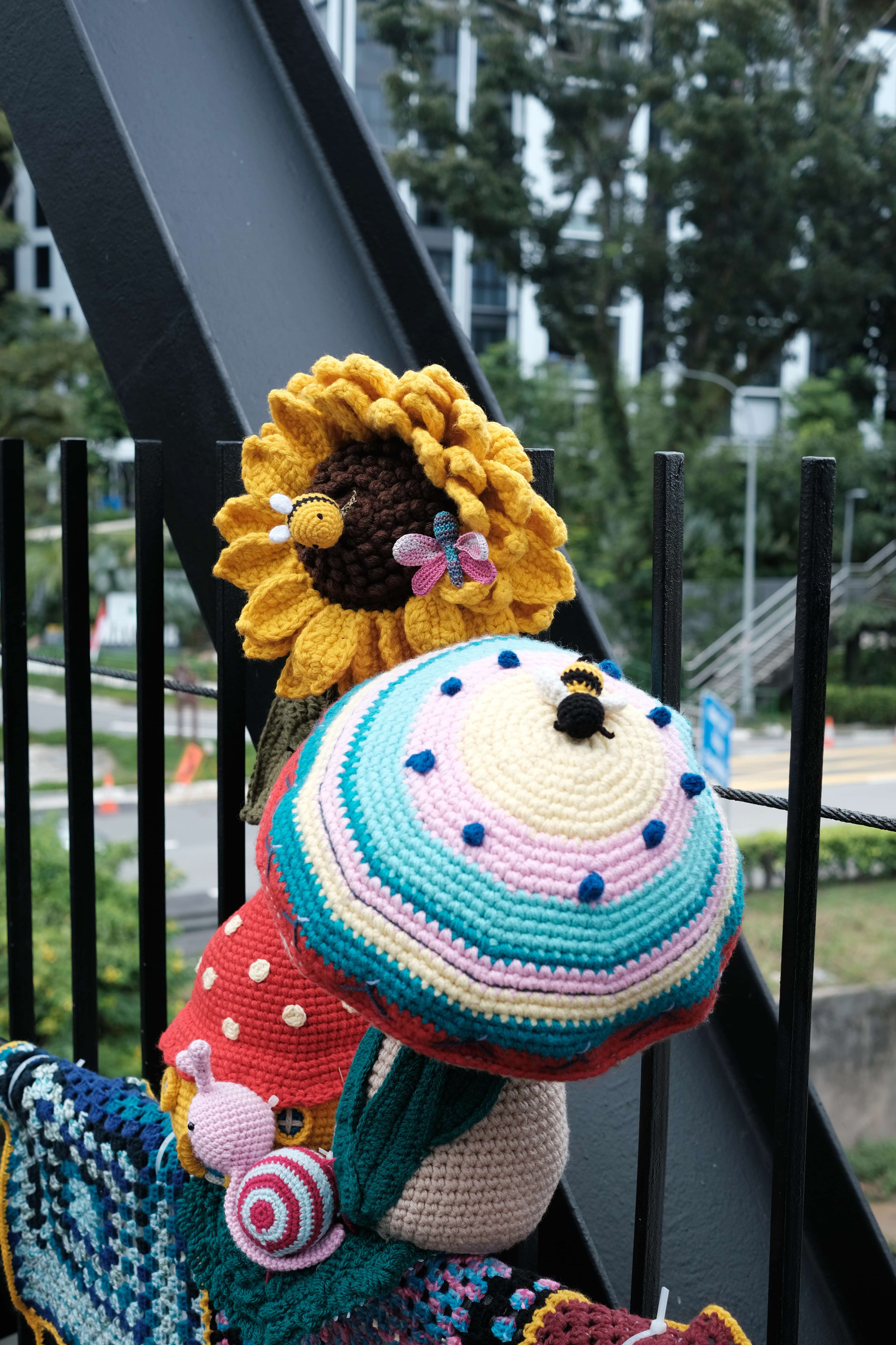 A close-up of crocheted amigurumi figures attached to a black metal railing outdoors. Prominently featured is a large yellow crocheted sunflower with a dark brown centre, adorned with a small crocheted bee and a pink and teal dragonfly. Below it sits a large crocheted mushroom with a colourful striped cap in teal, pink, and cream, decorated with blue spots and a small bee on top. To the left is a red polka-dotted fairy house, and a small pink crocheted snail sits at the base. Crocheted granny square blankets in blue and multicoloured patches are draped over the railing. Trees and a residential building are visible in the background.