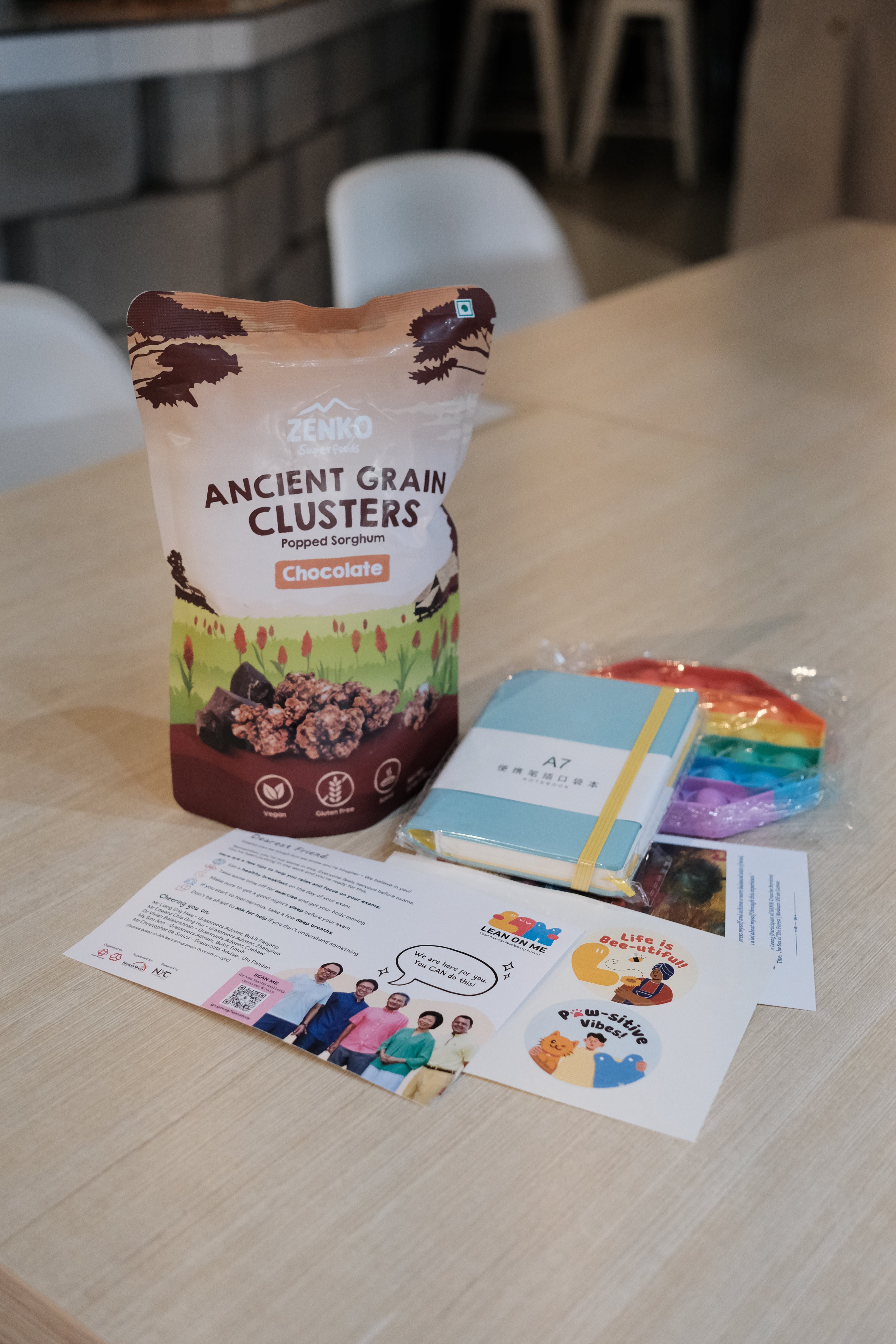 Zenko Ancient Grain Clusters chocolate snack bag beside a blue notebook, stickers, and a printed card on a wooden table.