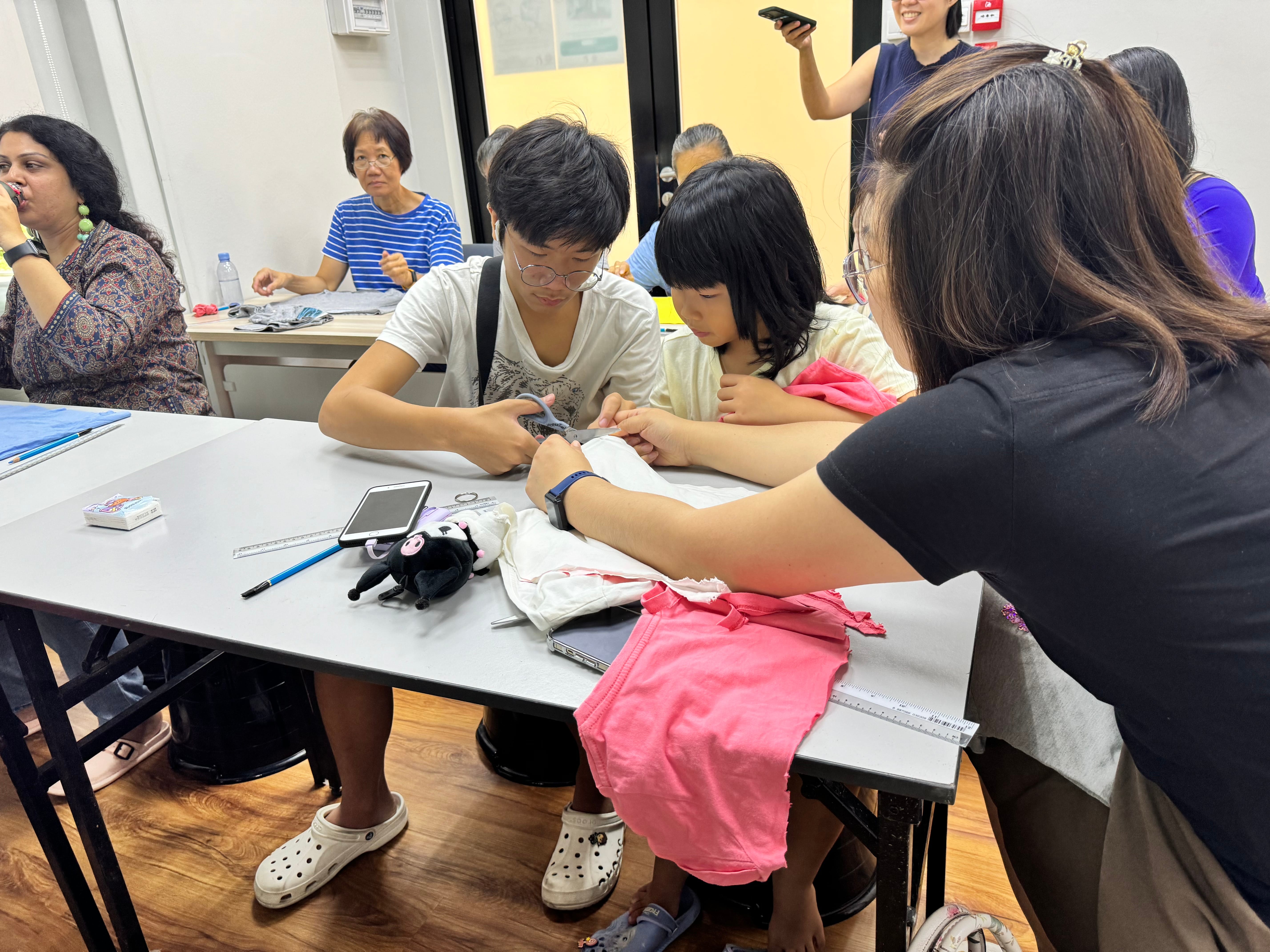 Two youths and a woman sit at a table sewing fabric, with phones, rulers, and cloth scattered in a classroom.