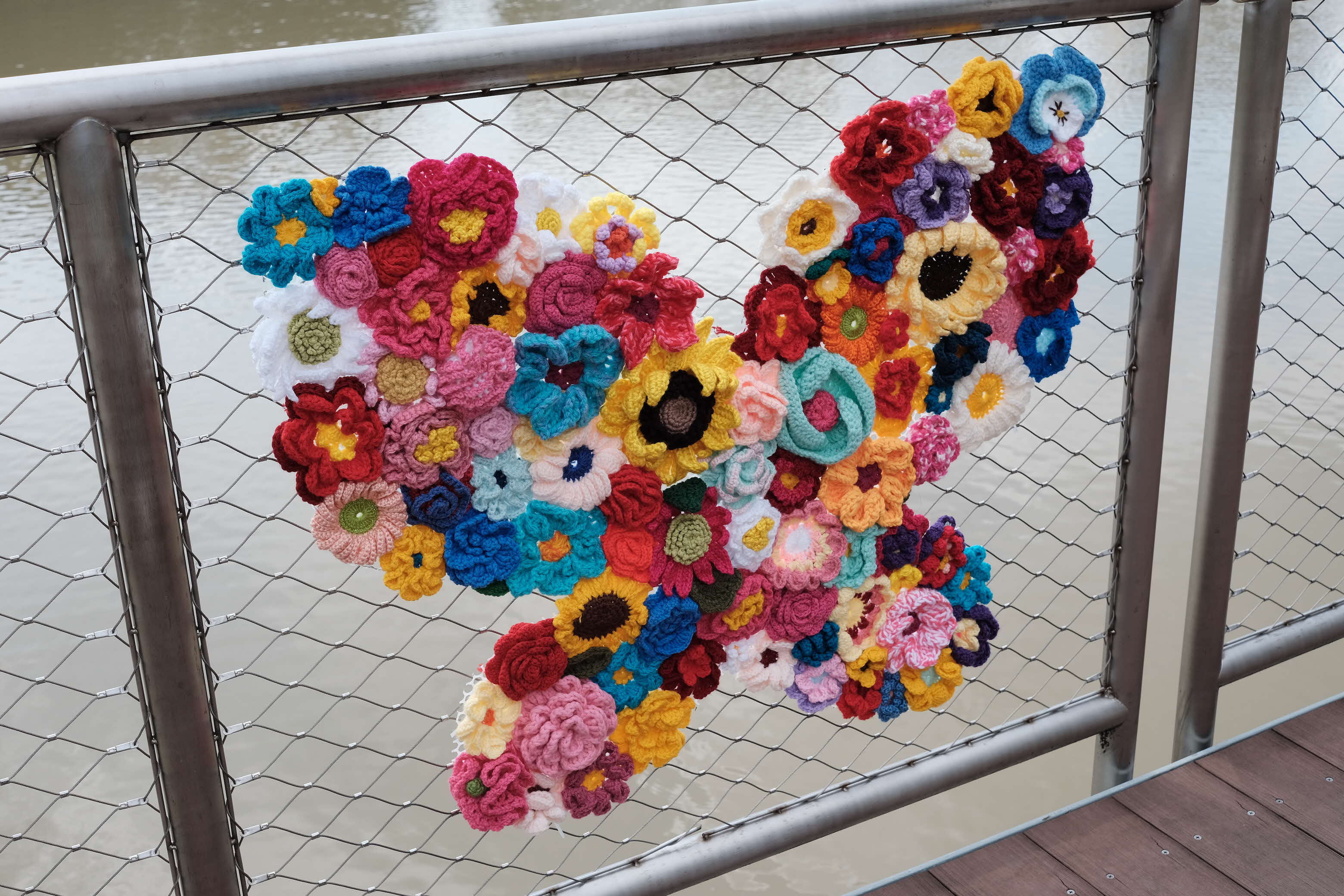A colourful crochet flower arrangement in a heart shape, attached to a metal mesh railing overlooking a pond.