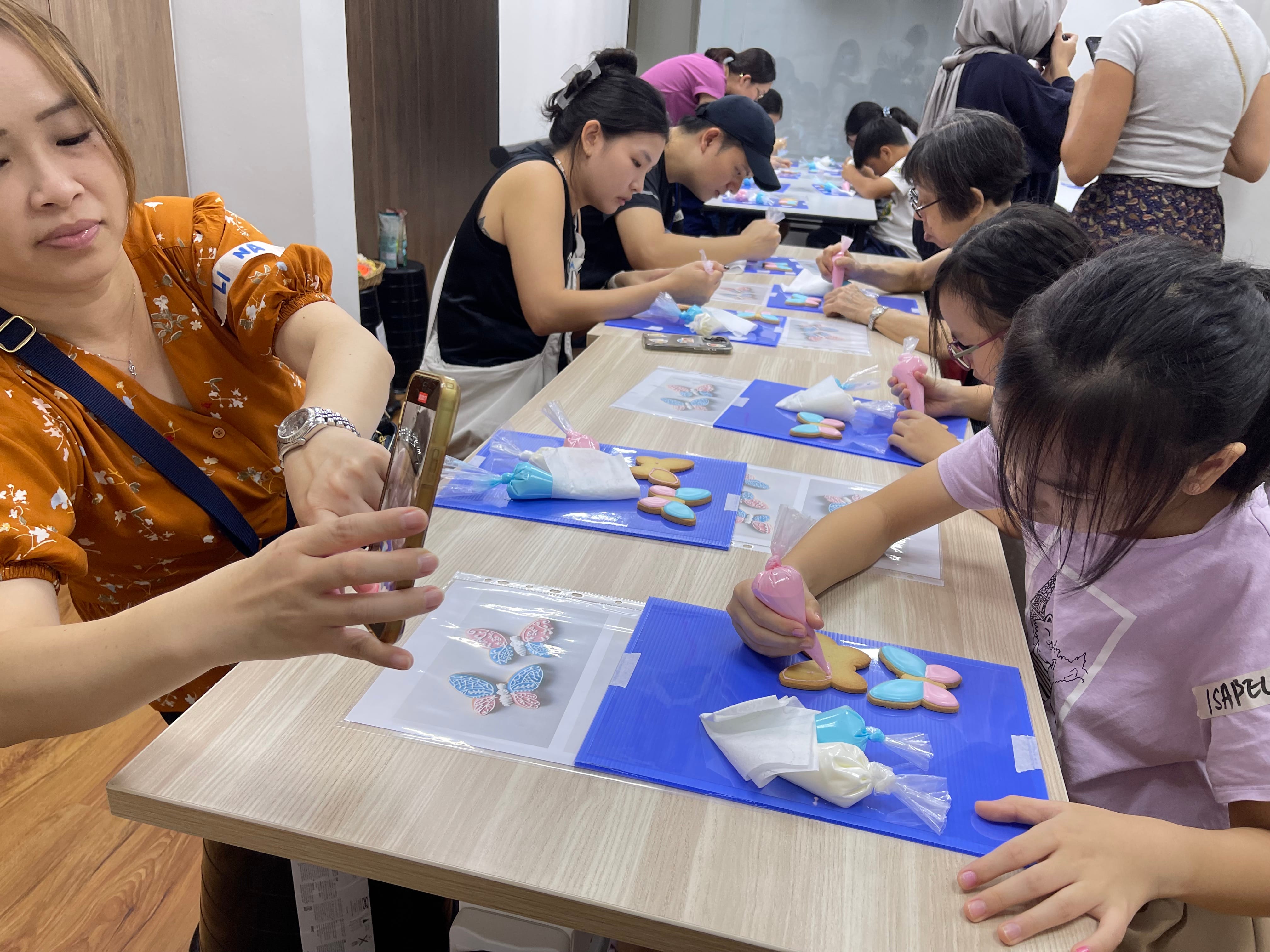 People at a long table decorating butterfly-shaped cookies with colorful icing bags during a cookie decorating class.