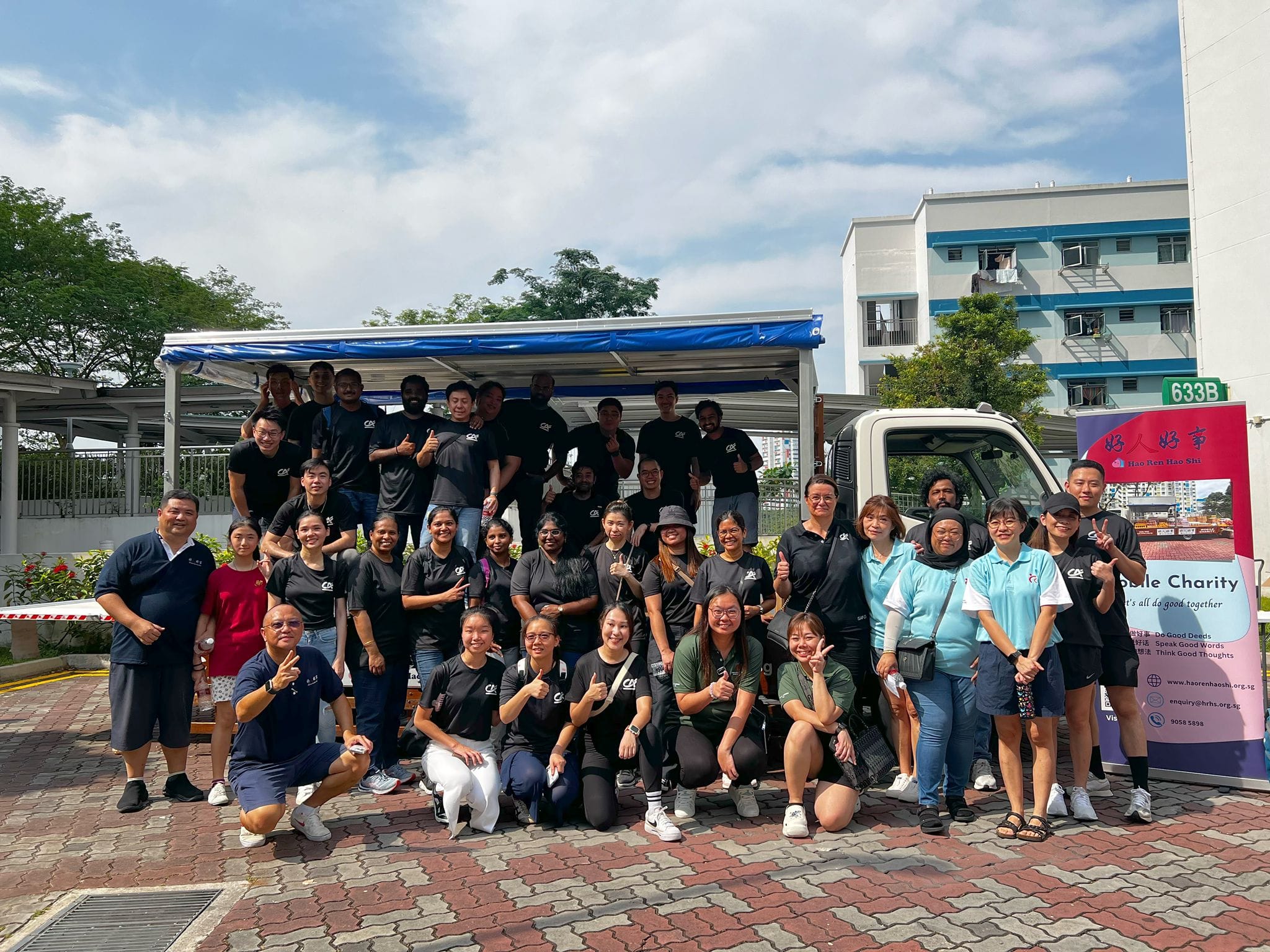 Large group of volunteers in black shirts posing outdoors near a truck and a Hao Ren Hao Shi banner in Singapore.