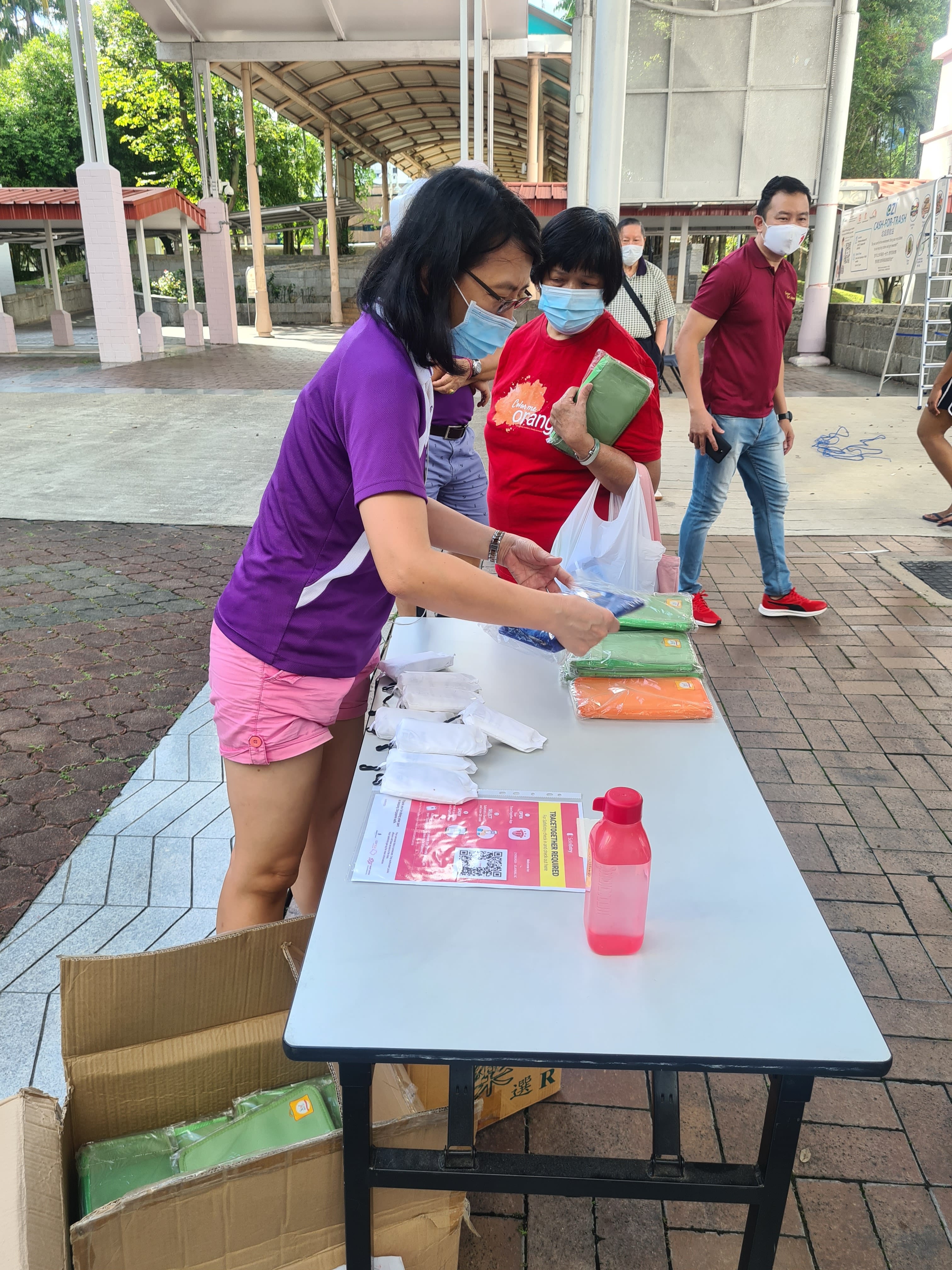 People wearing face masks stand at an outdoor table distributing folded eco bags as a reward for Cash for Trash.