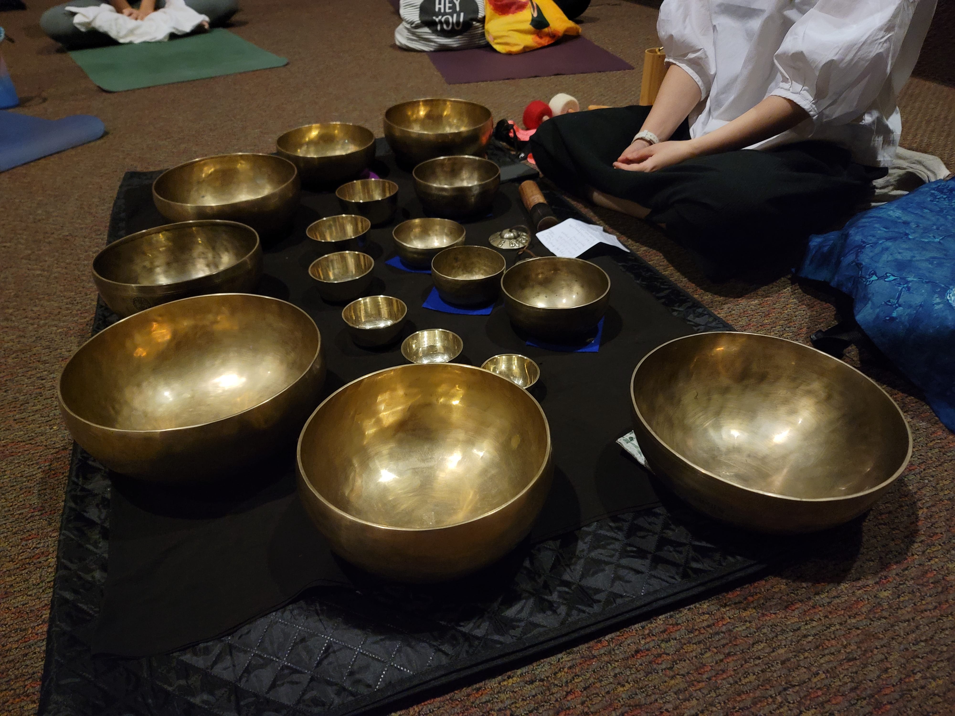 Multiple brass Tibetan singing bowls of varying sizes arranged on a black mat, with a person seated nearby.