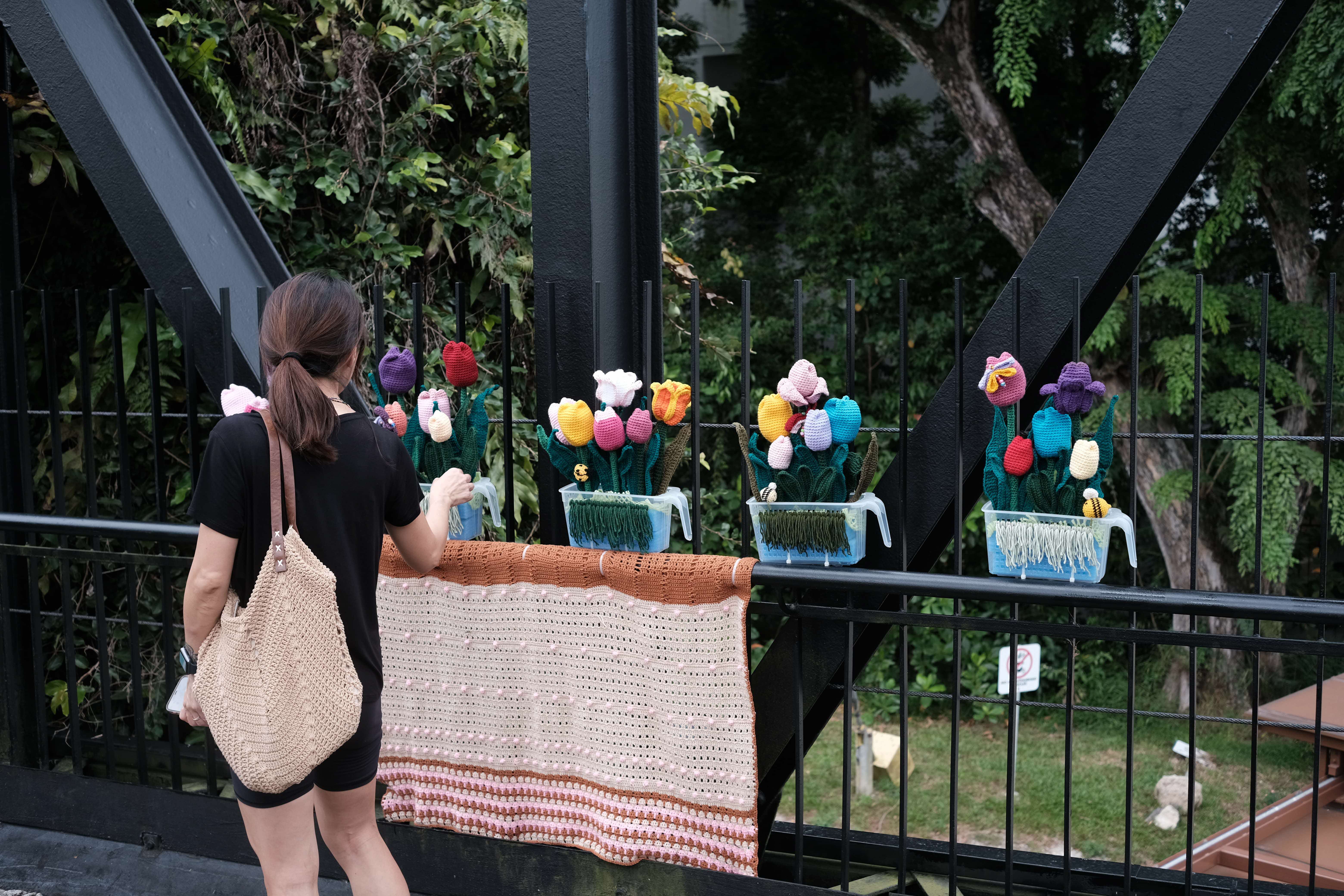 Woman with a woven bag looking at colorful crocheted flowers in planters on a black metal railing.