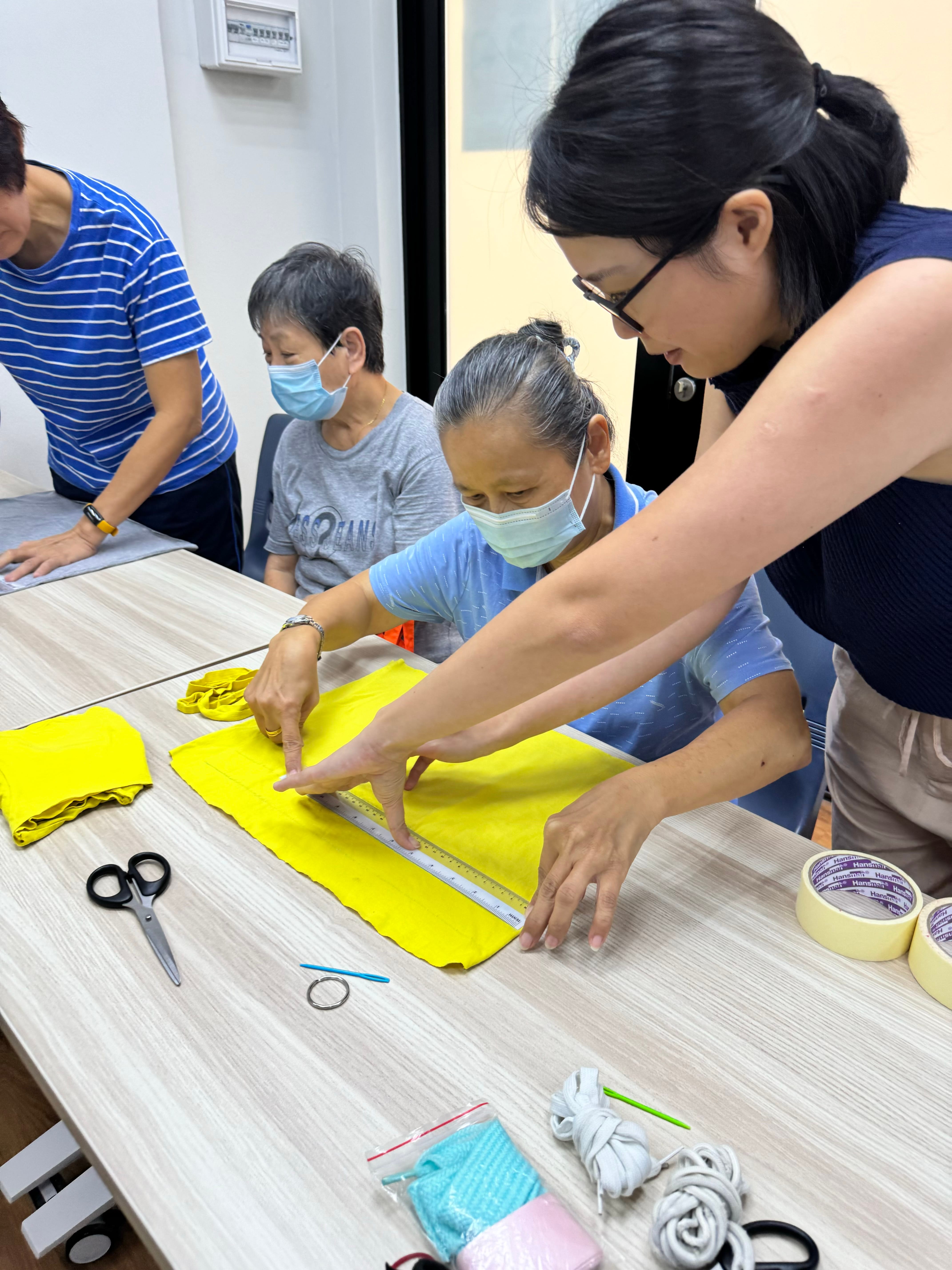A young woman guides two masked elderly women measuring yellow fabric on a table with scissors and tape nearby.