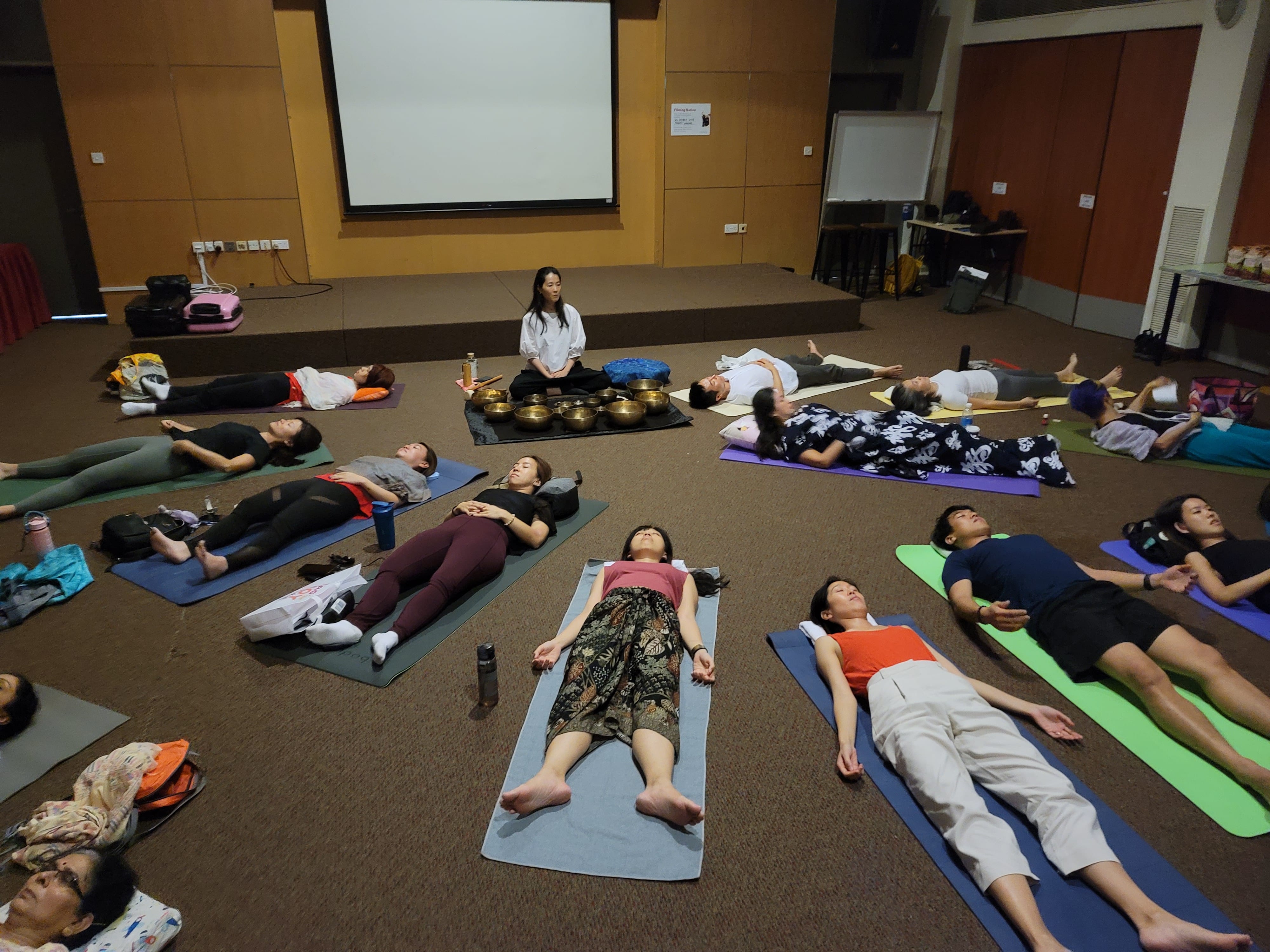 A group of people lying on yoga mats in a hall while an instructor sits with singing bowls nearby.