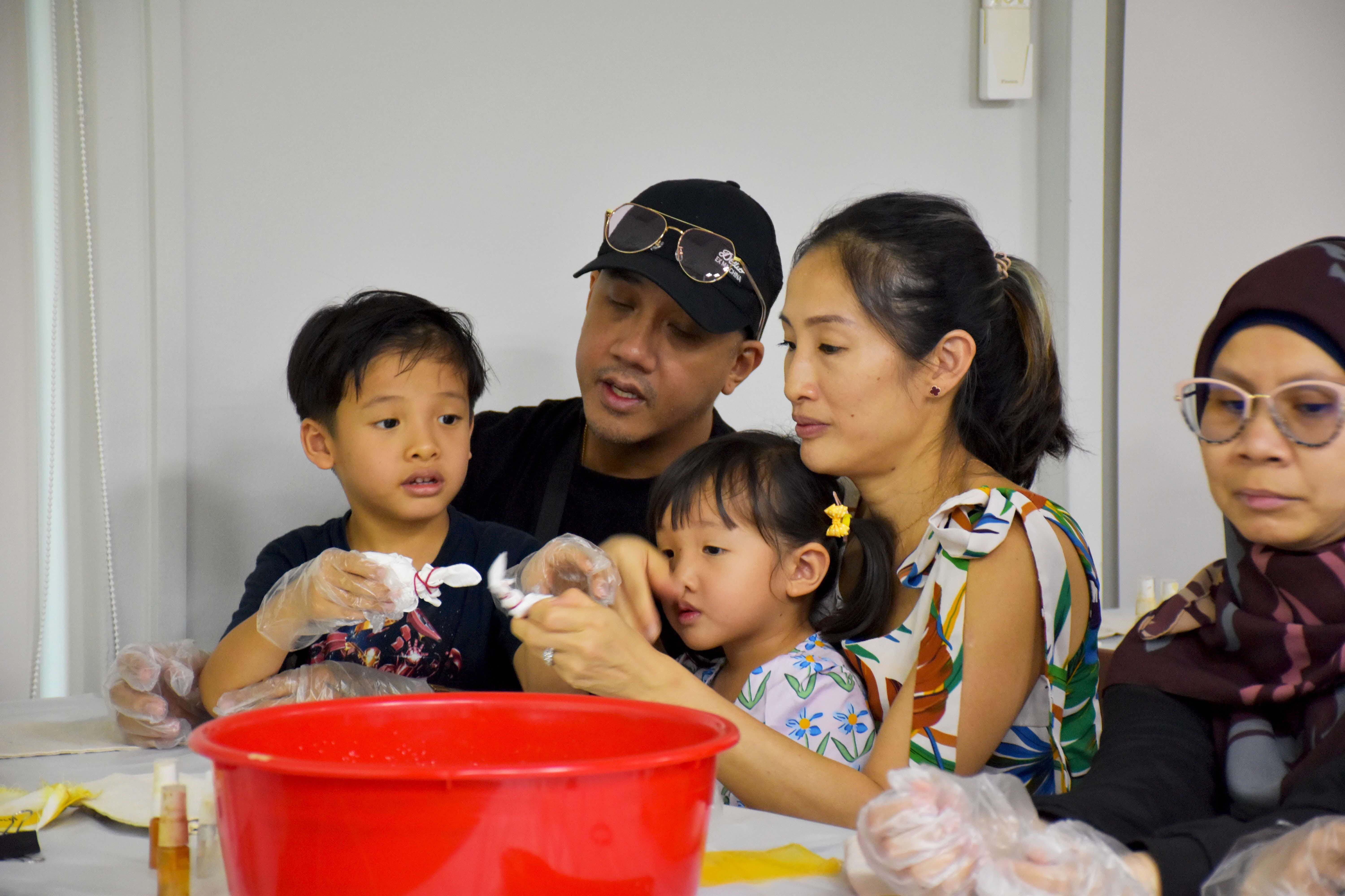 A family of four huddles closely together at a table during an indoor tie-dye craft workshop. A man in a black cap and sunglasses and a woman in a floral top lean in attentively as they help two young children — a boy and a toddler girl with yellow hair clips — handle white fabric with gloved hands. A large red basin sits on the table in the foreground, alongside dye bottles and yellow fabric. To the right, an older woman wearing a brown hijab and pink-framed glasses looks on. The group appears focused and engaged in the activity.