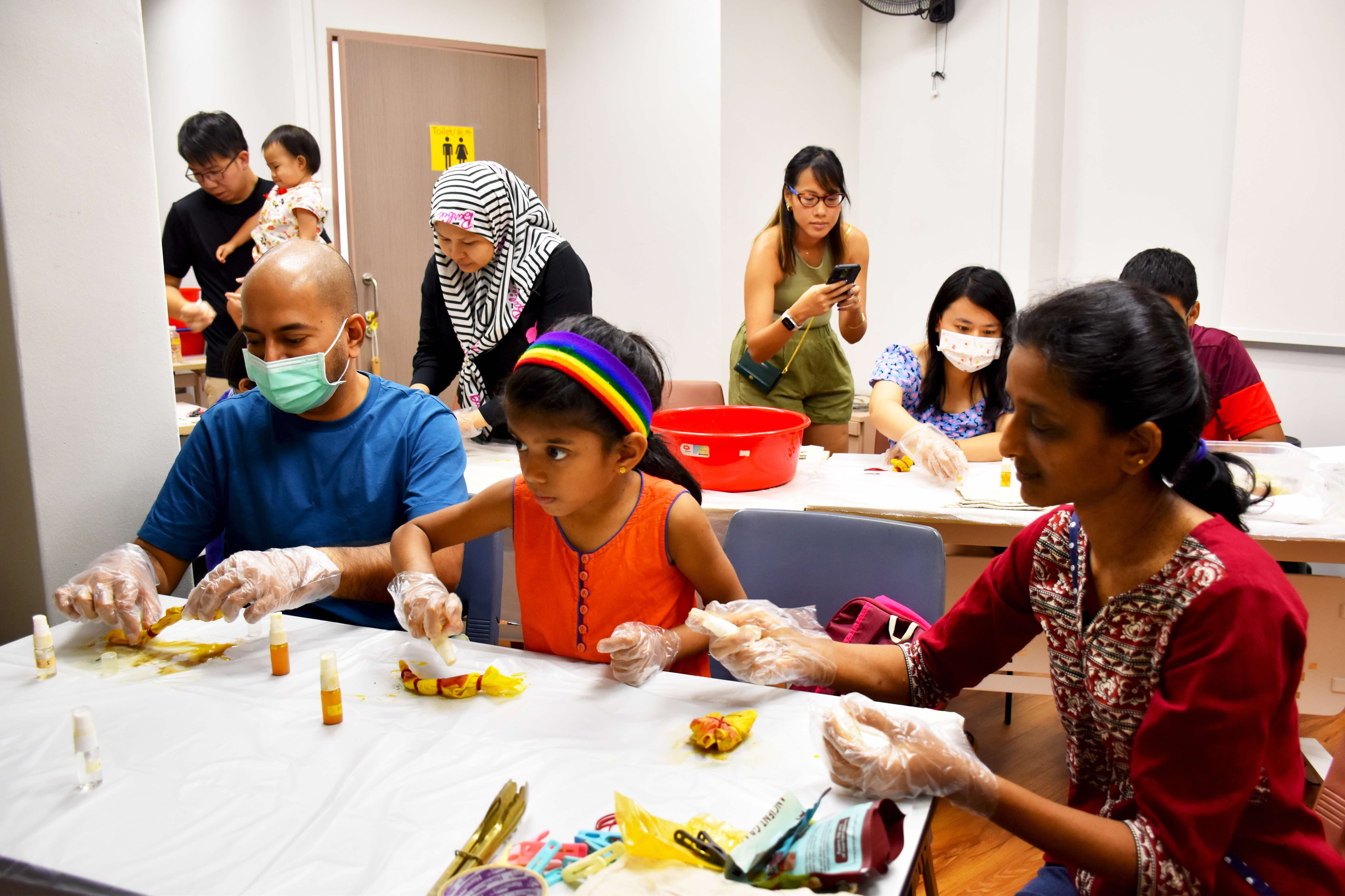 A diverse group of adults and children participate in an indoor tie-dye craft workshop. In the foreground, a man in a blue shirt and surgical mask, a young girl wearing a rainbow headband and orange dress, and a woman in a red patterned top work together at a table covered with white paper, applying yellow dye to fabric while wearing plastic gloves. Behind them, a woman in a black and white striped hijab assists at the same table, while other participants work at tables in the background. Dye bottles, rubber bands, and fabric pieces are visible on the tables. A red basin sits on one of the tables in the middle of the room.