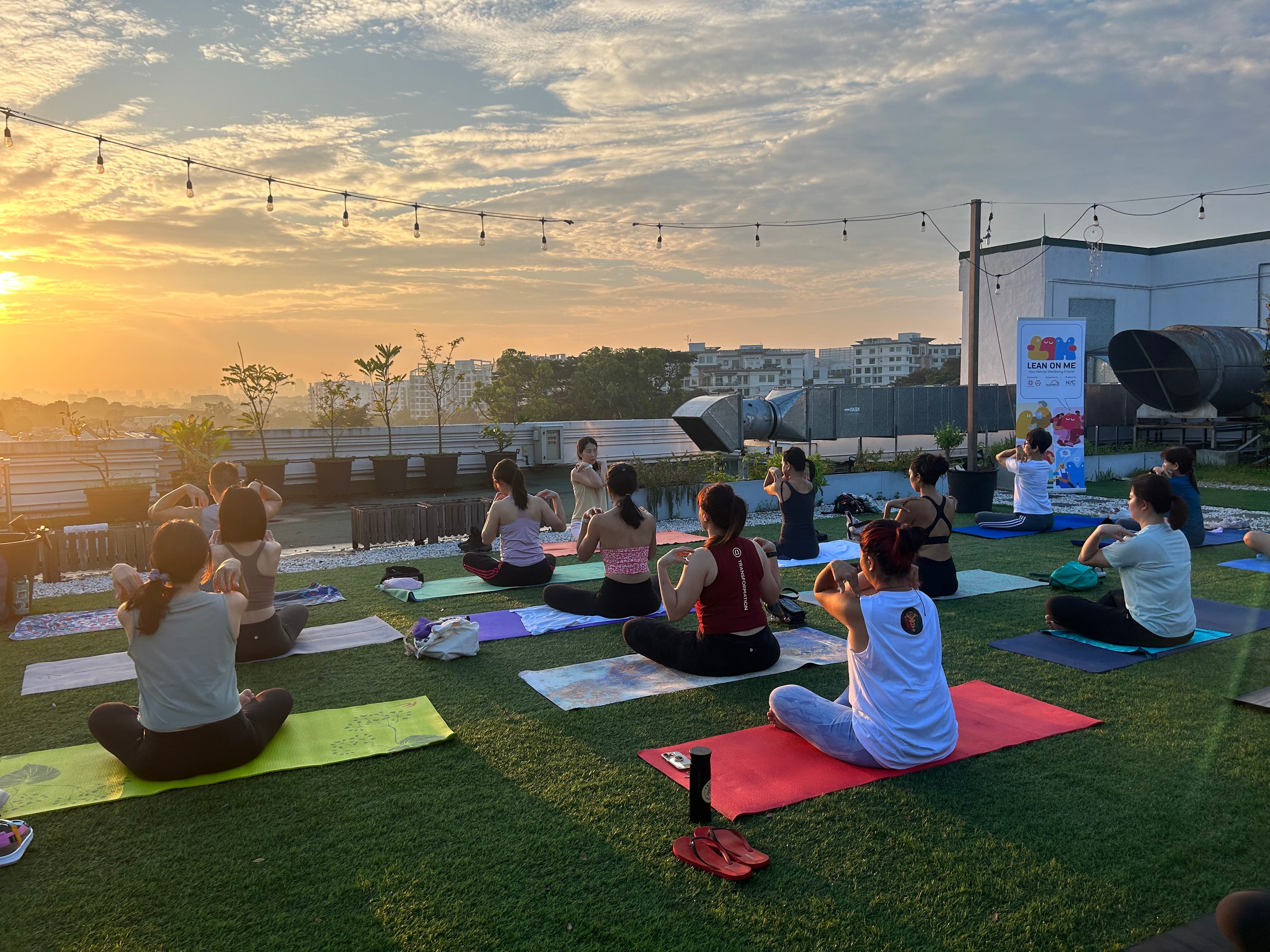 Group of people doing yoga on colorful mats on a rooftop at sunset with string lights overhead.
