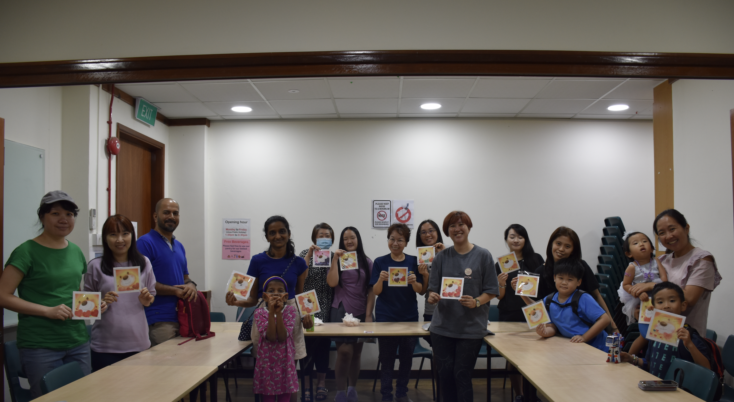 Group of adults and children in a classroom holding handmade cards, standing by U-shaped tables.