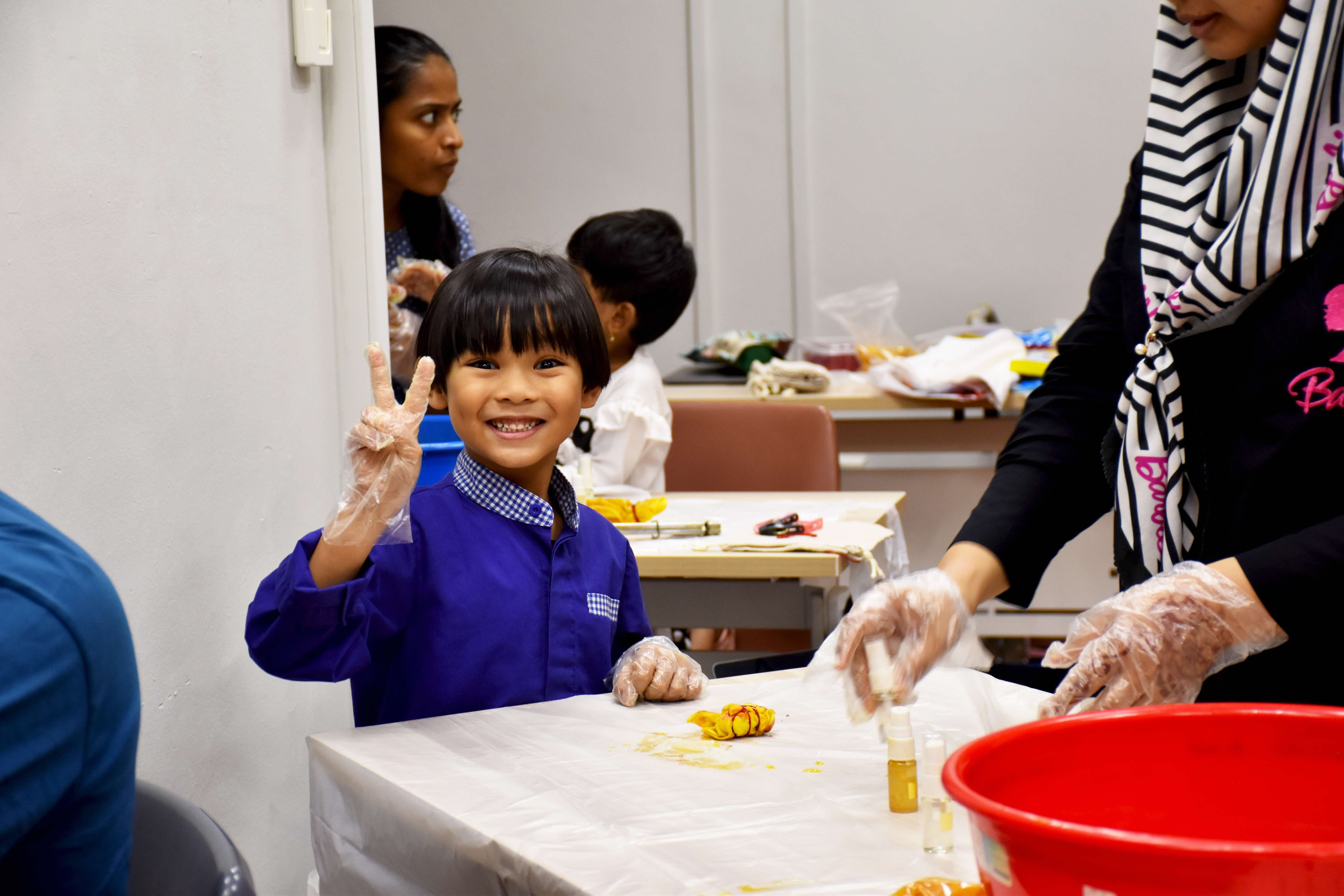 A young child in a blue shirt grins at the camera and flashes a peace sign with a gloved hand during an indoor tie-dye craft workshop. A small piece of yellow-dyed fabric sits on the white paper-covered table in front of them. To the right, an adult wearing a black and white striped hijab assists at the table, also wearing plastic gloves and holding a dye bottle. A red basin is visible at the edge of the table. In the background, other children and an adult are seated at tables engaged in the same activity.