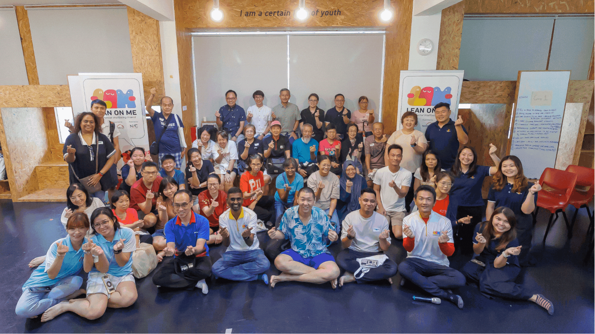 Large group in a room posing and smiling, many making finger-heart gestures; posters and chairs behind.