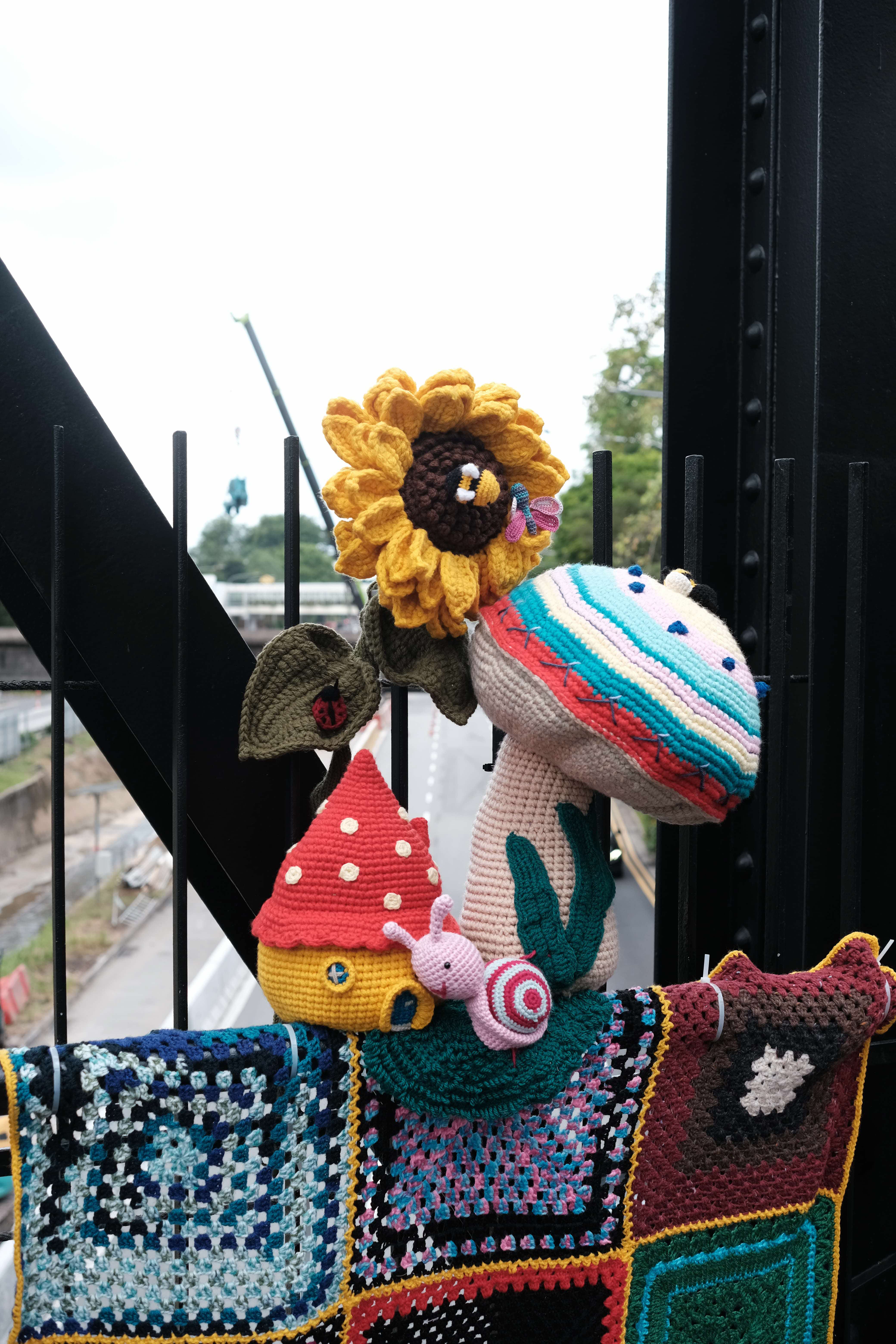 A colourful collection of crocheted amigurumi figures and blankets displayed on a metal railing outdoors. The arrangement features a large yellow sunflower with a small bee, a multicoloured striped mushroom, a red and yellow fairy house, a pink snail, a green leaf with a ladybird, and teal plant stems. Below the figures, crocheted granny square blankets in blue, black, and multicoloured patches are draped over the railing. A road and construction site are visible in the background.