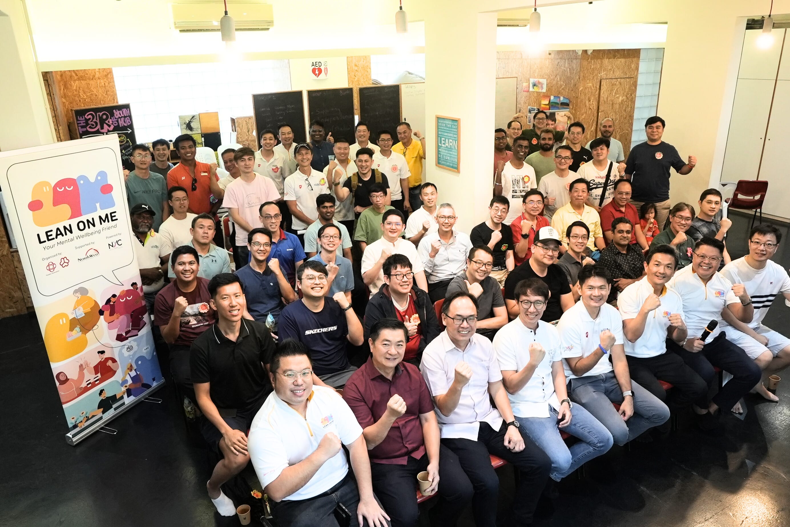 Large group of people posing with raised fists indoors next to a Lean On Me mental wellbeing banner.