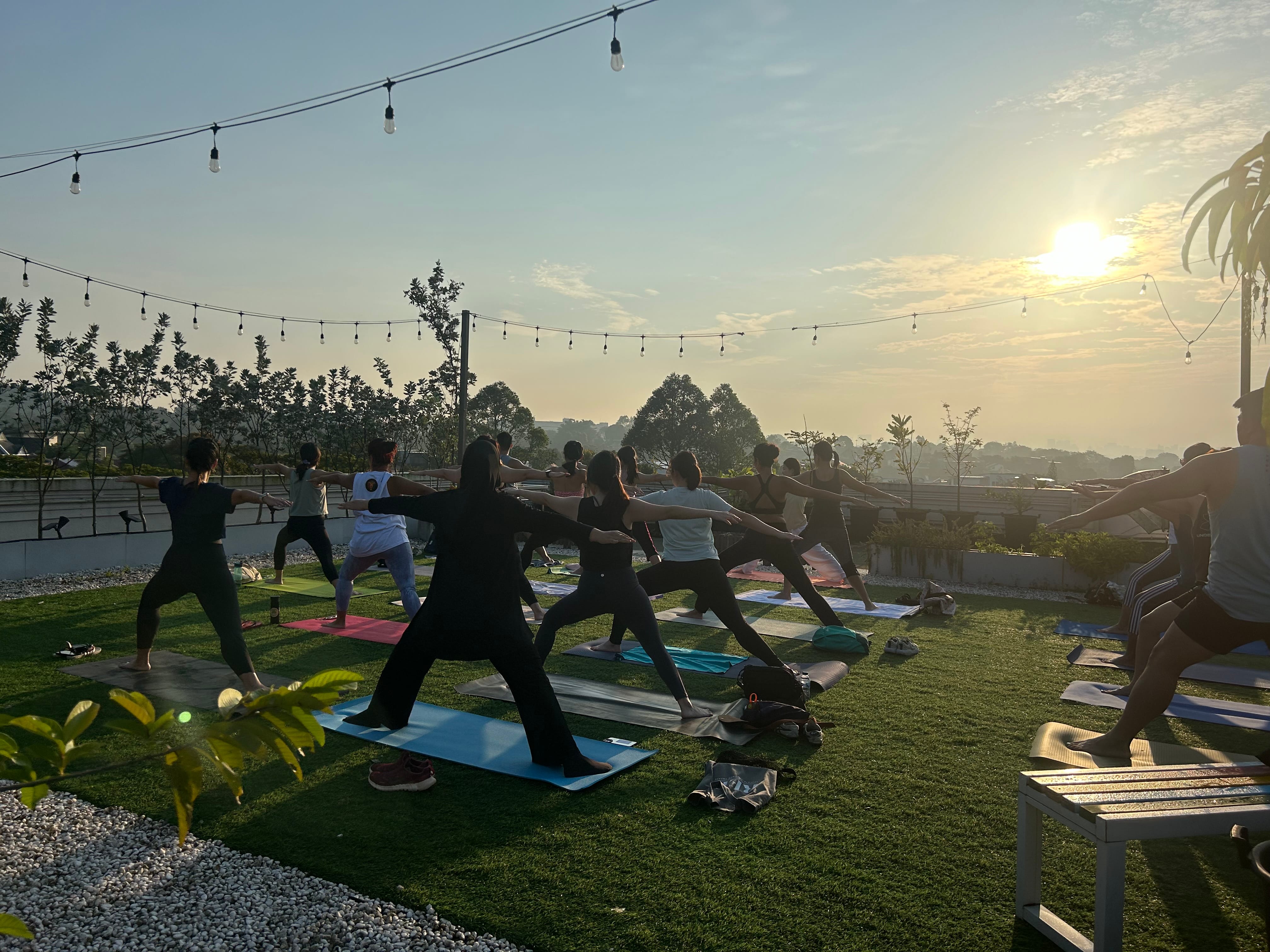 Group of people doing warrior yoga pose on mats outdoors at sunset, with string lights overhead.