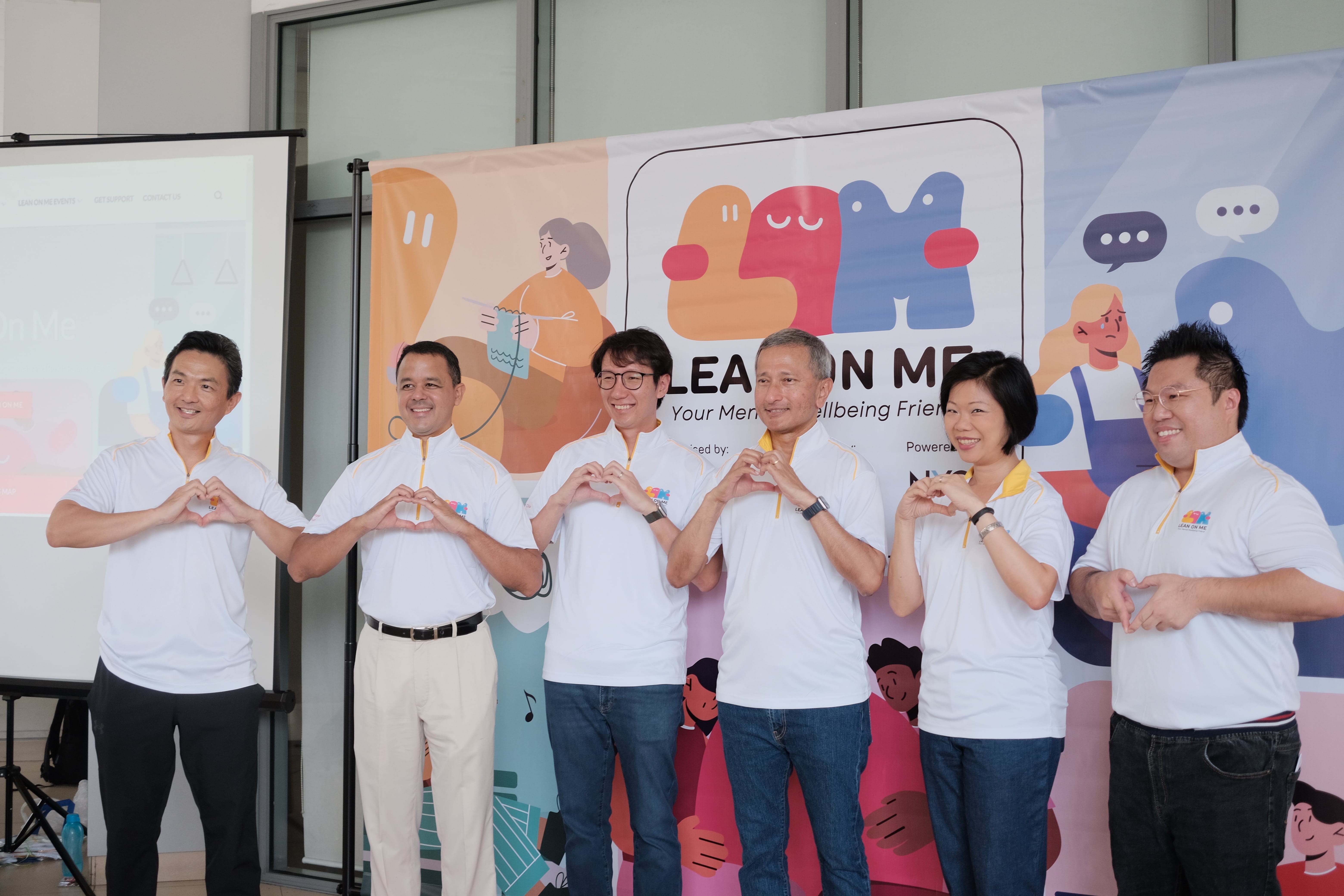 Six people in white polo shirts make heart shapes with their hands in front of a colorful Lean On Me banner.