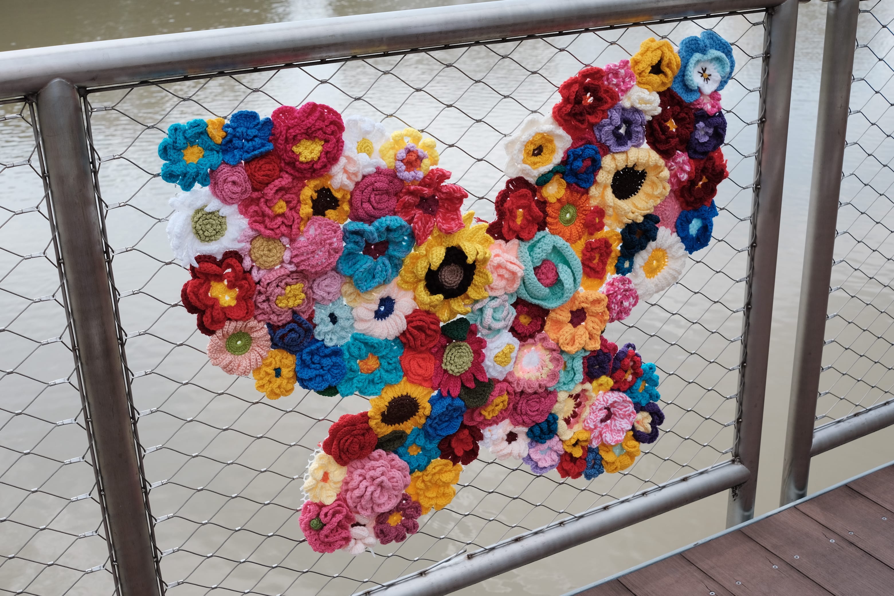Colorful crocheted flowers form a butterfly shape attached to a metal mesh railing by the water.