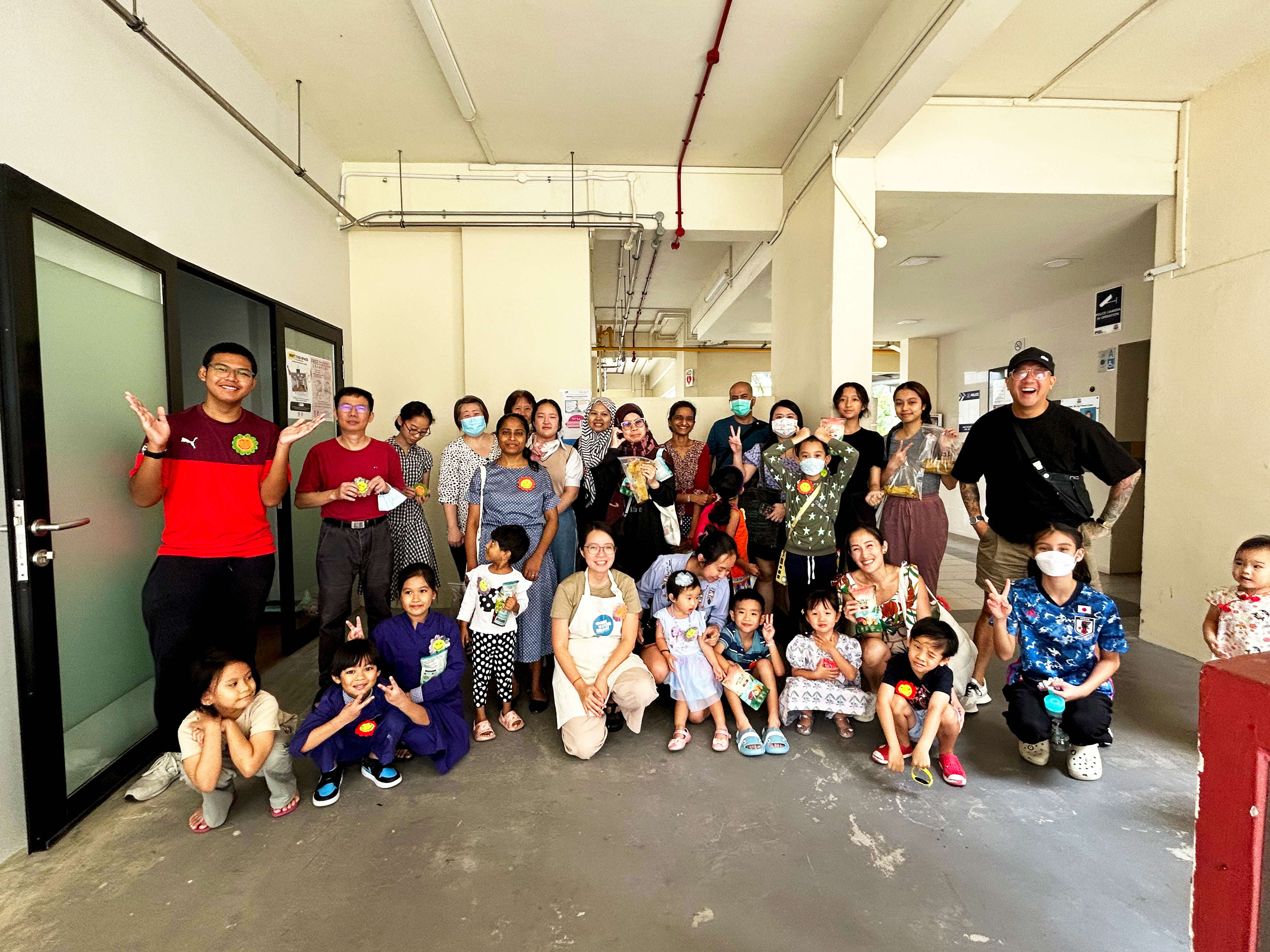 A large diverse group of adults and children pose together for a group photo in the corridor of a public housing block. The group of approximately 25 people spans multiple generations and ethnicities, with several participants holding up packaged craft items, likely tie-dyed fabric from a workshop. Many are smiling and flashing peace signs at the camera. One woman in the centre wears a white apron. The group includes young children crouching in the front row, adults standing in the middle and back rows, and a toddler standing to the far right. Some participants are wearing face masks.