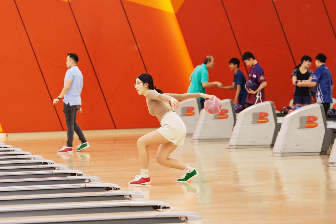 A young woman in a skirt releases a pink bowling ball at a colorful orange bowling alley with other players nearby.