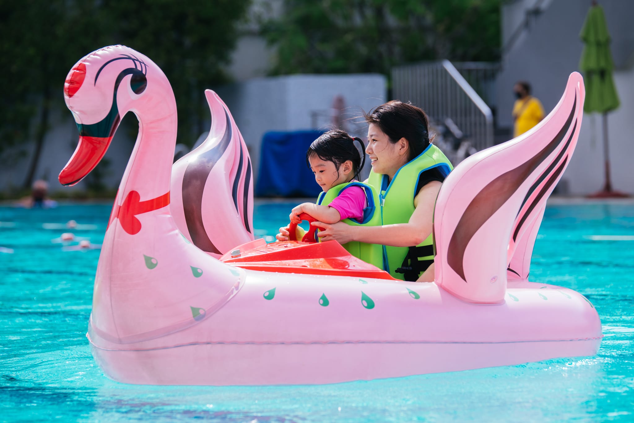 A woman and young child wearing green life vests sit on a pink inflatable swan float in a pool.