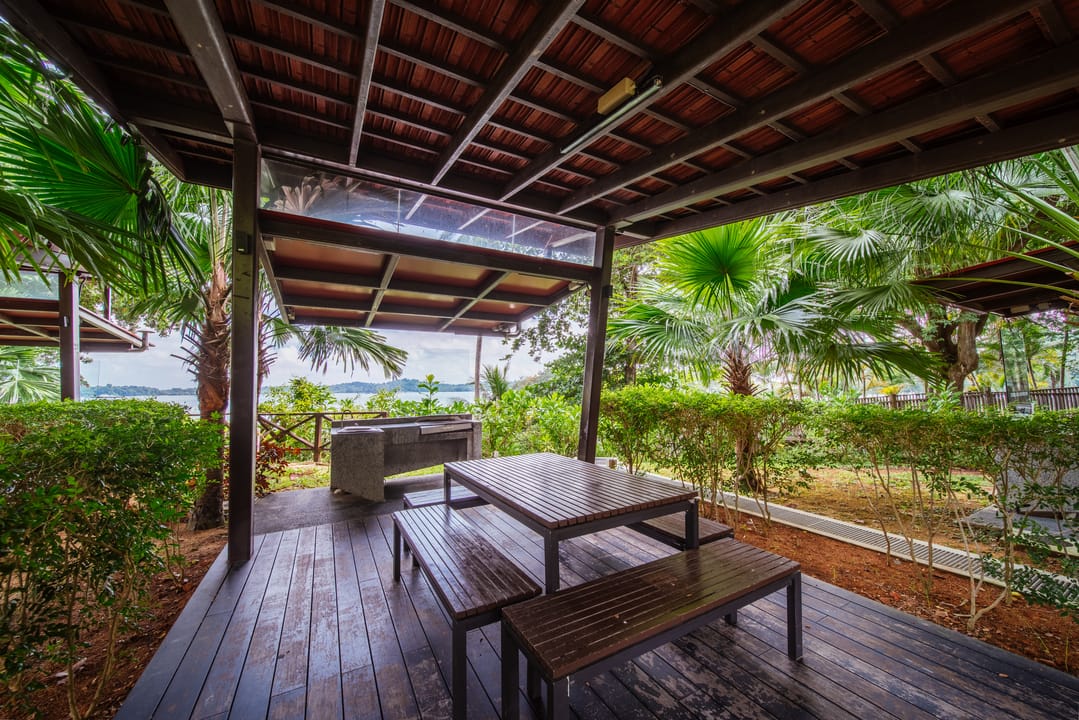 Covered wooden deck with benches and a table, surrounded by tropical plants with a water view in the background.
