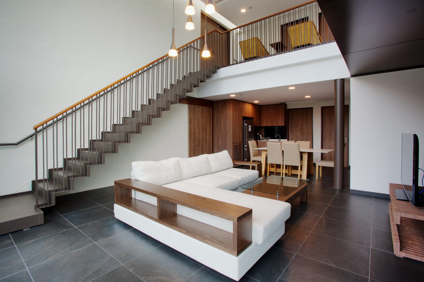 Modern two-story living space with white sofa, wood coffee table, staircase with metal railing, and open kitchen behind.