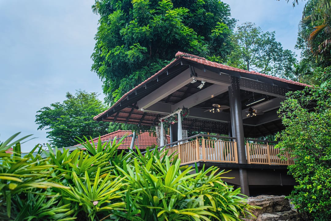An open-sided pavilion with a red tiled roof and wooden deck, surrounded by lush tropical greenery and tall trees.