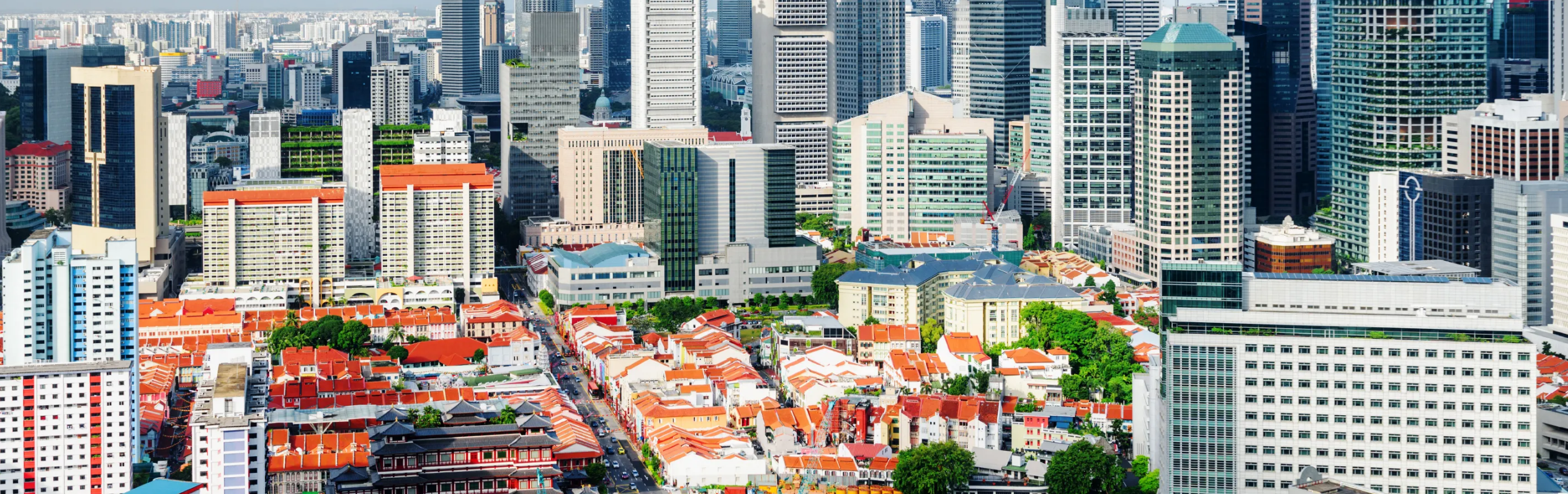 Dense city skyline with tall office towers behind rows of low buildings with red roofs and green trees.