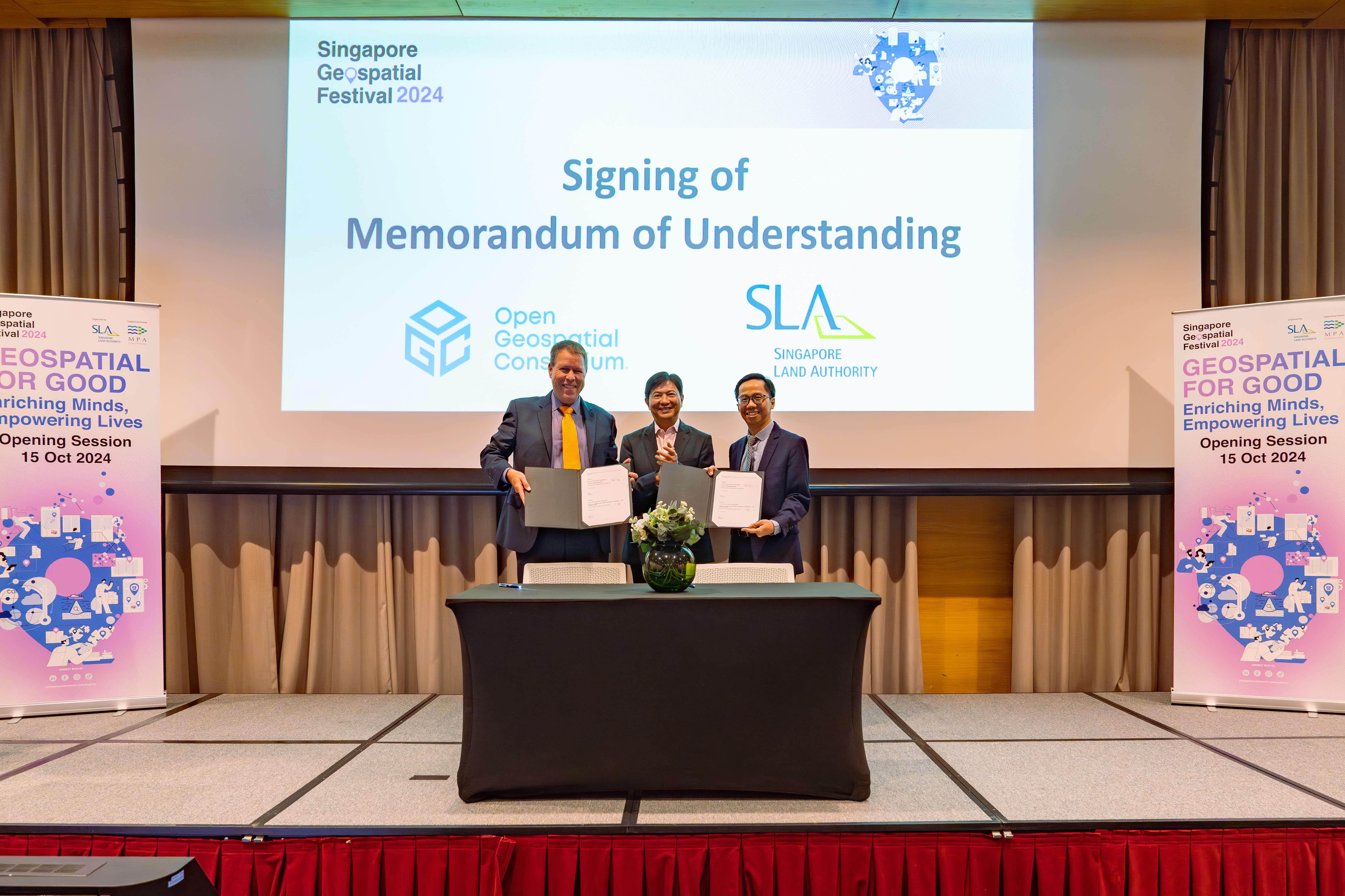 Three men on stage hold signed MOU documents at Singapore Geospatial Festival 2024 between OGC and SLA.