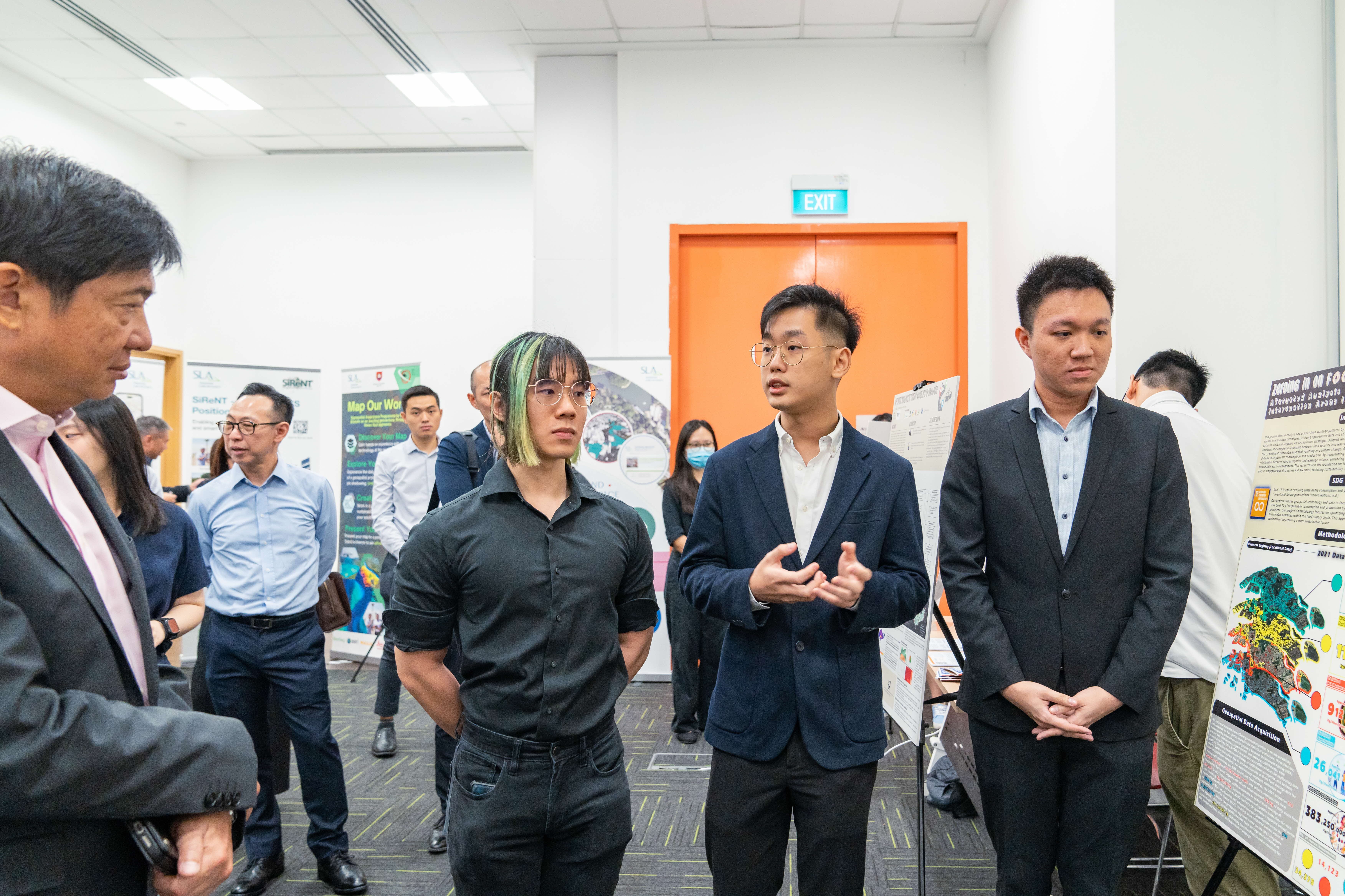 Several people stand in a room near research posters, with one young man in a suit gesturing while speaking to others.