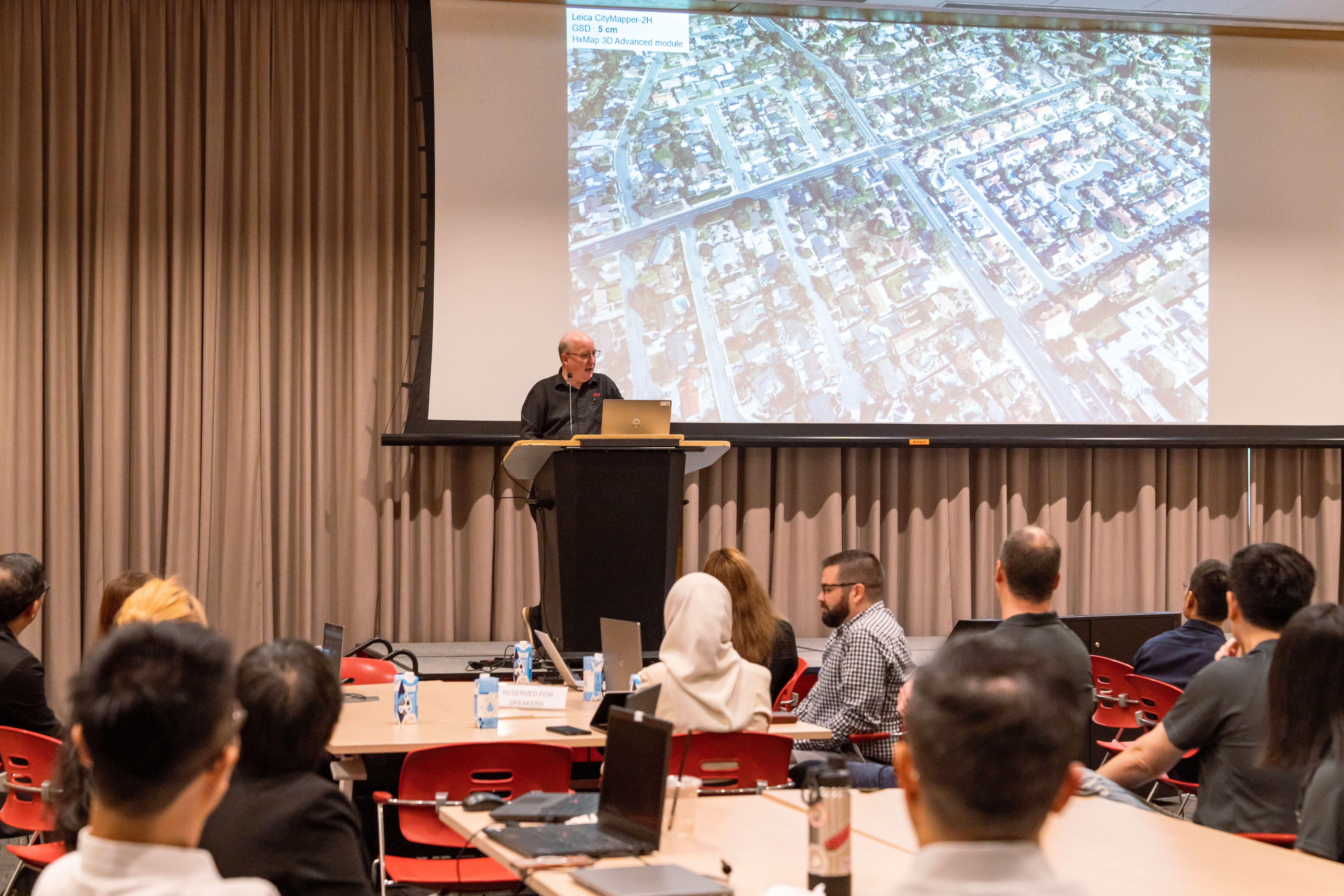 A man presents at a podium to a seated audience, with an aerial city map projected on a screen behind him.