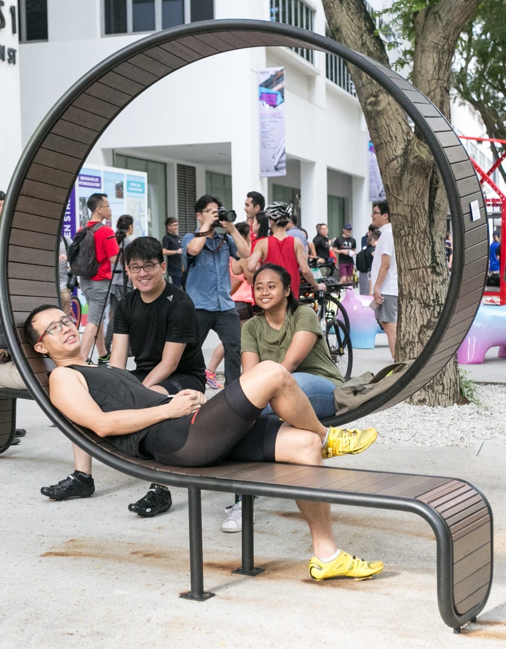 Man reclines on a rounded bench as people look on and a photographer takes pictures.