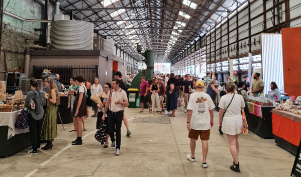 Indoor market scene with many people browsing stalls under a high, metal-framed roof.