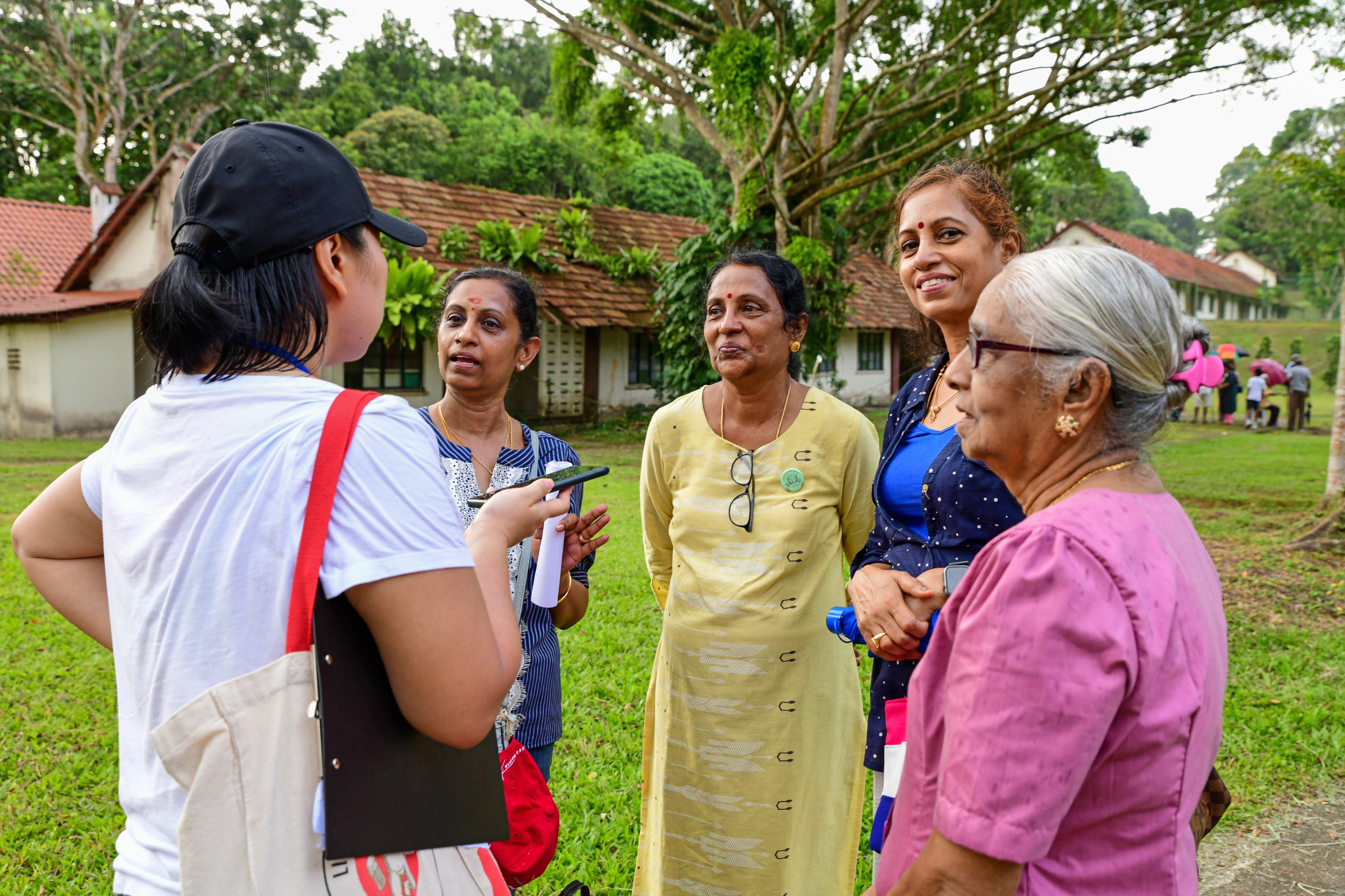 A person interviewing four women outside in a park. They're standing on a lawn in front of a building.