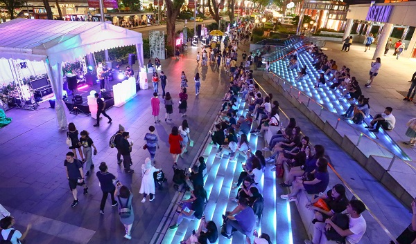 Nighttime scene of a crowd watching a band perform on an outdoor stage. Illuminated steps provide seating.