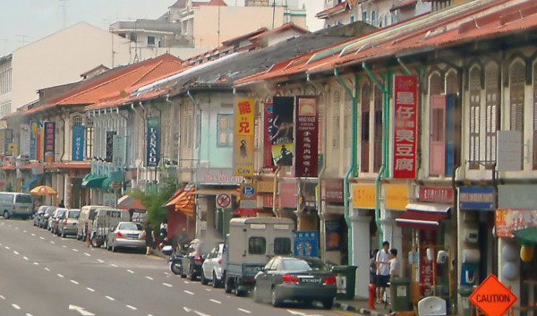 Street view in Singapore. Traditional shophouses line the road with parked cars and signs.
