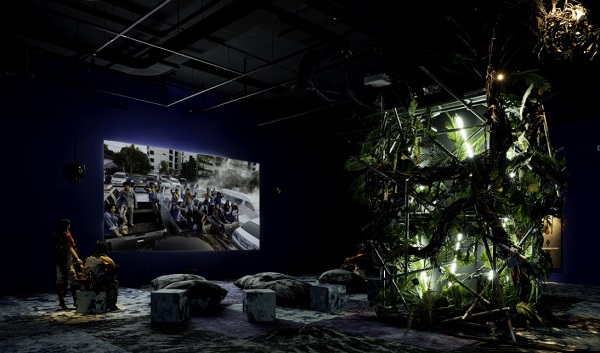 Art installation in a dark room, featuring a projected video and a metal cube covered in plants. People are viewing the video.