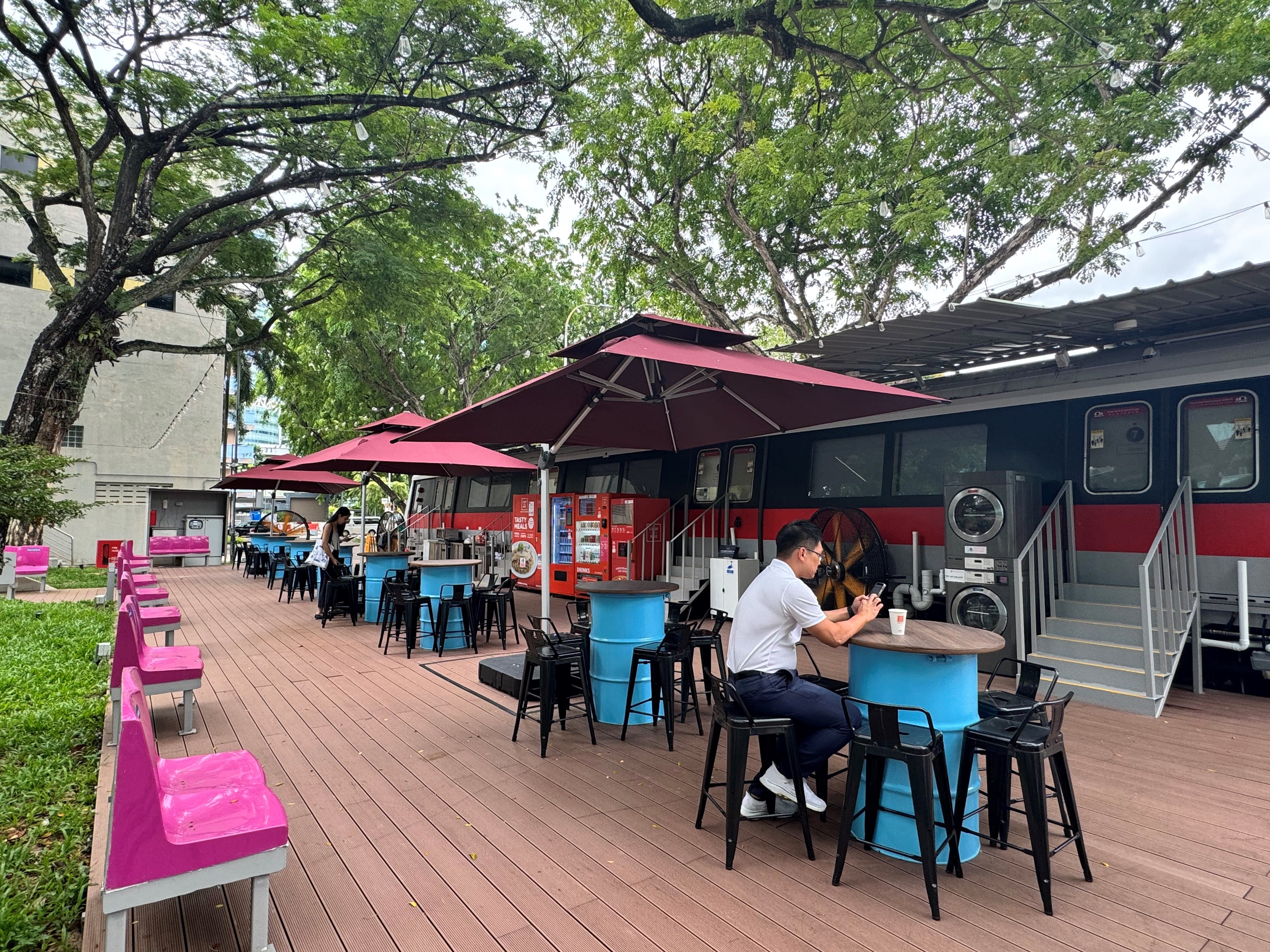 Outdoor cafe with tables made from blue barrels, black chairs, and red umbrellas. A person uses a phone at one table.