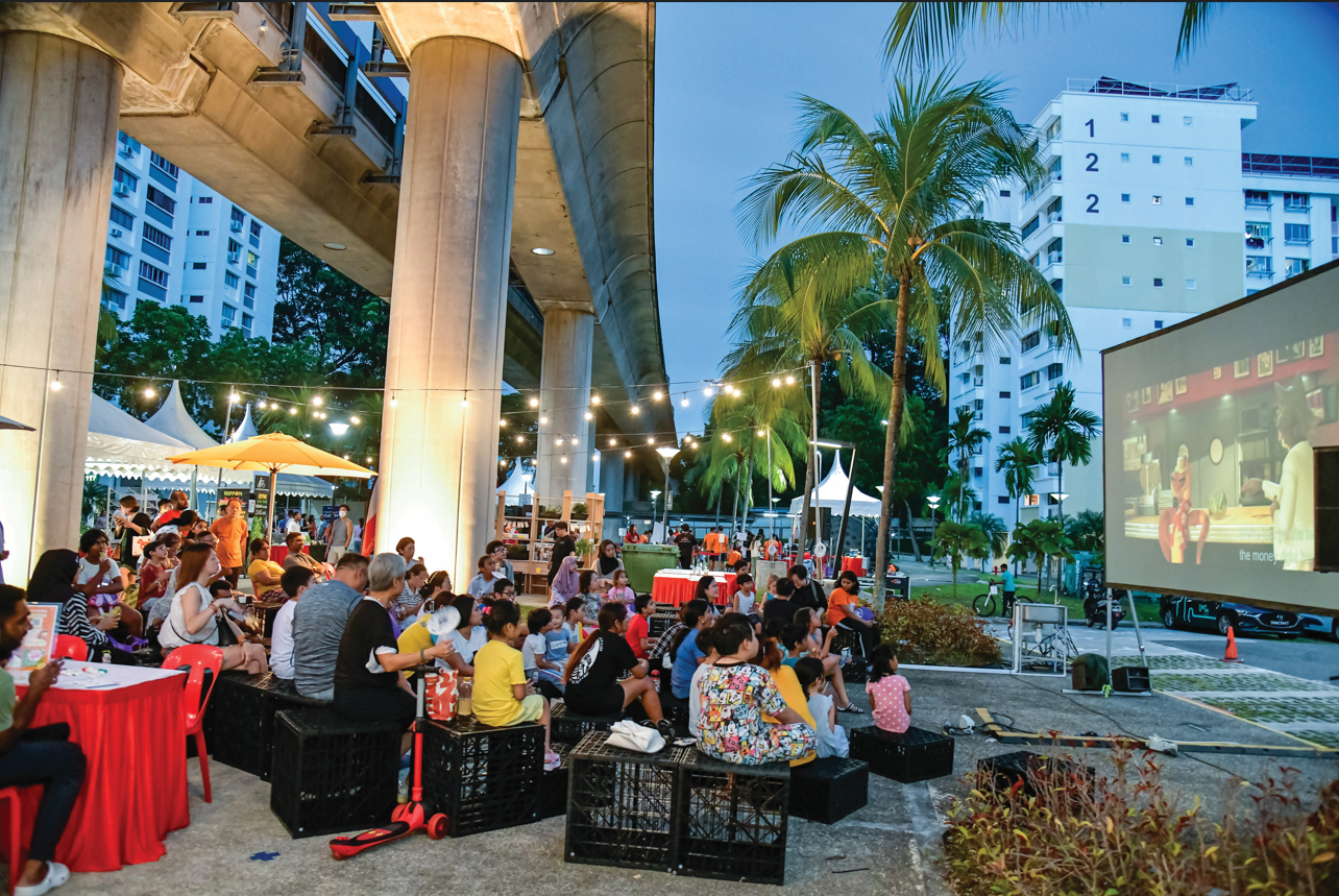 Outdoor movie screening with diverse crowd, under string lights and an elevated train track, in a park with palm trees, at dusk.