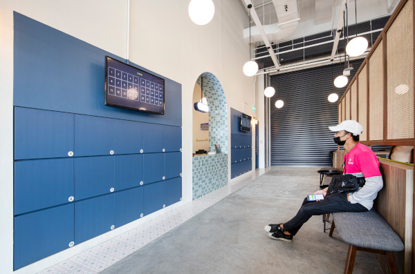 Interior view of a delivery hub with blue lockers, monitors, seating, and a masked person waiting.