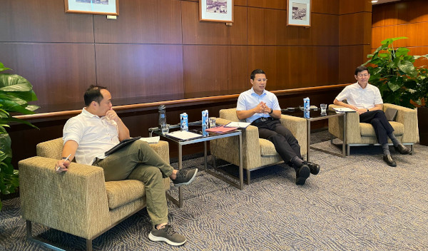 Three men sit in armchairs around small tables in a room with paneled walls.